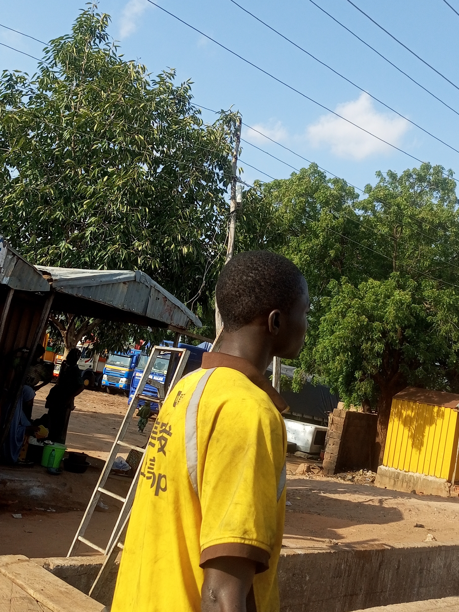 In Gombe, Nigeria, a young Black man wearing a bright yellow shirt with reflective stripes and some dark lettering is standing outdoors, facing away from the camera and looking to his right. He has short, dark, curly hair. The man is positioned in the foreground, with a rough concrete structure visible in the lower part of the frame. Behind him, a row of parked trucks is visible. The trucks appear to be a mix of colors, including blue, white, and yellow. A silver ladder leans against a makeshift shelter made of corrugated metal. Several trees with lush green foliage are scattered throughout the background, and a clear blue sky with a few wispy clouds is overhead. There are power lines crisscrossing the sky. The ground is a dusty, reddish-brown color. To the right of the man, a bright yellow corrugated metal structure stands on a concrete base, casting a distinct shadow on the ground. A person in a blue garment and another in darker clothing are partially visible in the shade of the shelter, near the ladder. A green bucket and a black basin are on the ground near them. The overall scene suggests an outdoor marketplace or industrial area.