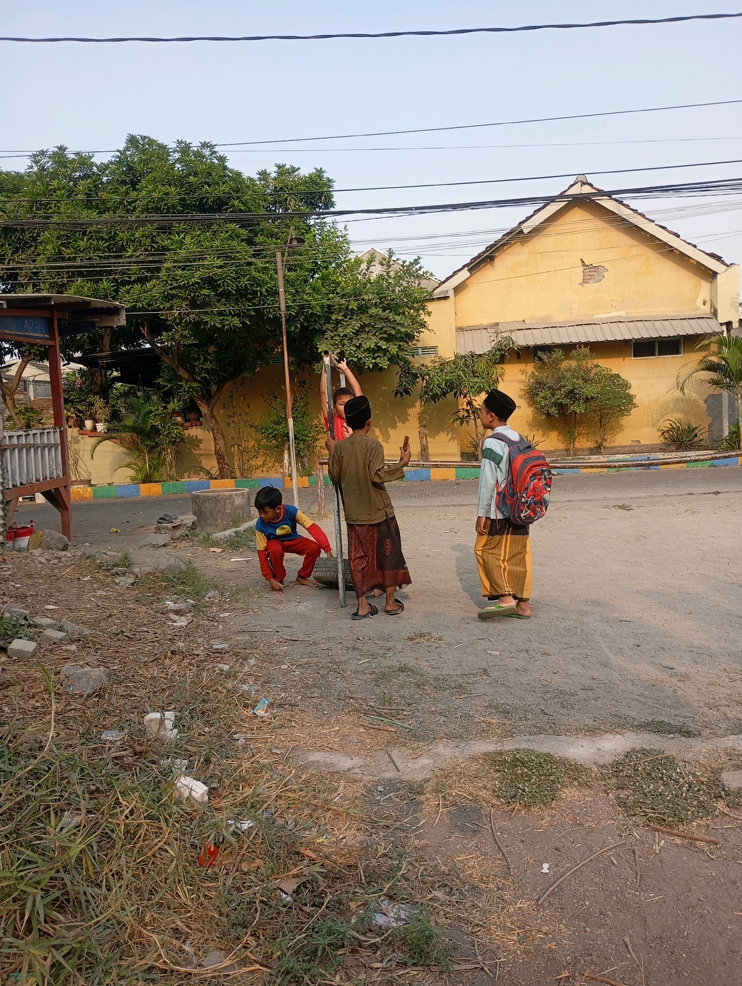 In Modong, Indonesia, three young boys are observed in an outdoor, possibly rural or suburban, setting. The scene appears to be a dusty, unpaved area with scattered debris and vegetation. One boy, wearing a blue and yellow shirt with red pants, is crouched down, seemingly interacting with a tire. Another boy, in a green and white shirt and yellow pants, stands facing away from the camera, carrying a backpack with a red and floral design, and wearing a peci (a cap common in Indonesia). He is standing near a metal pole. A third boy, wearing a brown long-sleeved shirt and patterned sarong, is also near the pole, holding what appears to be a mobile phone. Behind them, a yellow building with a peaked roof and visible damage to its facade is visible. Trees and other vegetation are present throughout the image, and power lines stretch across the sky. The ground is a mix of dirt, gravel, and dry grass. The overall impression is a casual, everyday scene of children playing or interacting outdoors.
