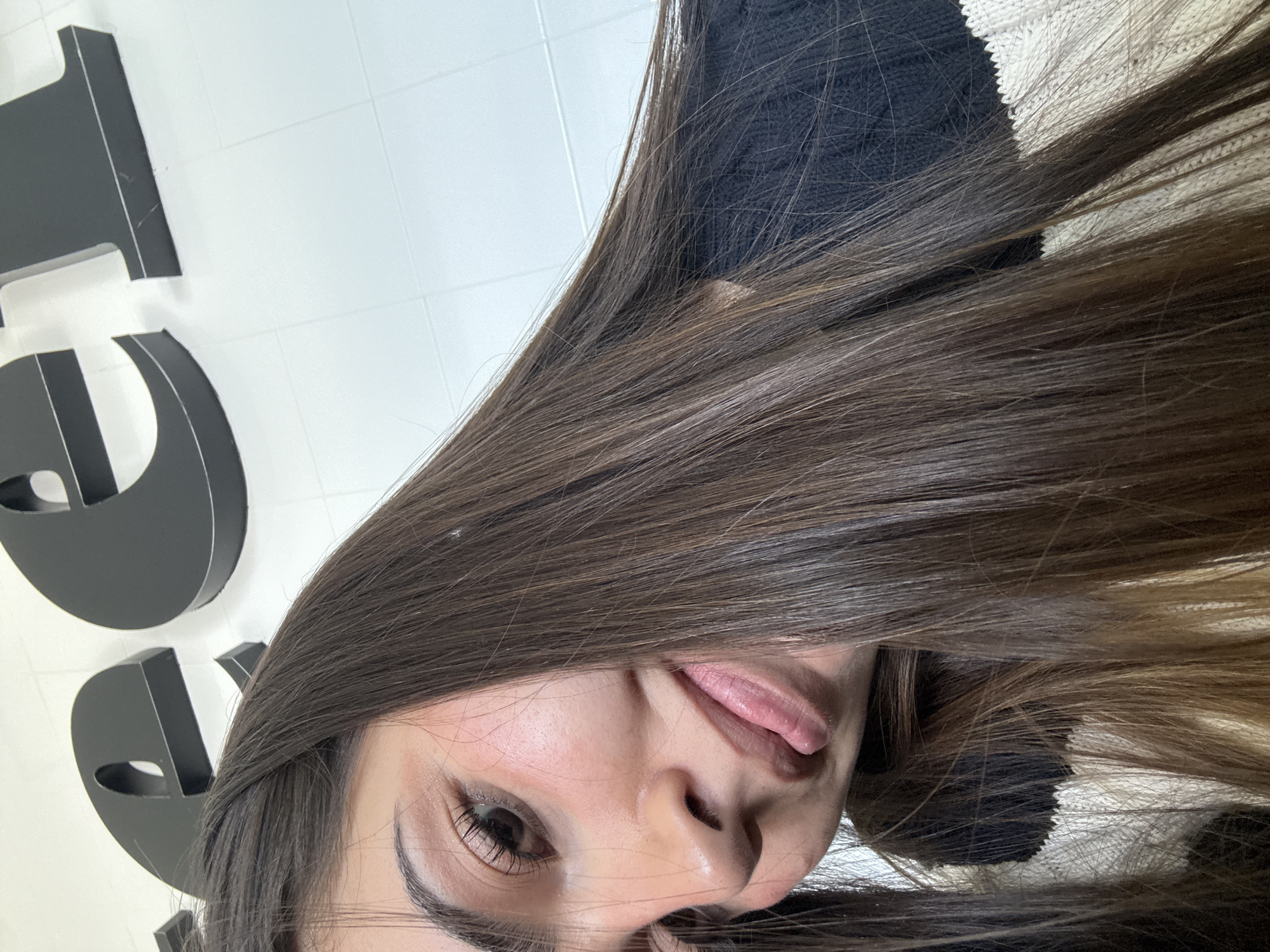 A young woman with long brown hair is positioned diagonally in the frame, with the upper left of the image showing partial black lettering on a white tiled wall. Her face is partially obscured by her hair, revealing one eye and part of her nose and mouth, with her tongue slightly protruding. She is wearing a dark-colored top with a portion of a white sweater visible. The location is possibly in San Bernardino Tlaxcalancingo, Mexico, based on the prompt instructions, although there is no visual confirmation of this.