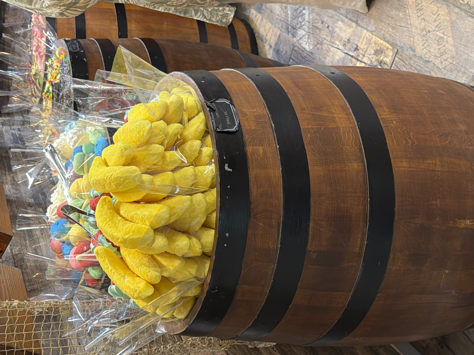 A close-up, eye-level shot shows a wooden barrel overflowing with bright yellow, banana-shaped marshmallows. The barrel has two black metal bands. A small black label is attached to the front of the barrel, reading "Marshmallows HALAL." Behind this barrel, another similar barrel is visible, also filled with various colorful candies. The background consists of wooden planks and a rough-textured wooden wall. The scene appears to be inside a candy store.