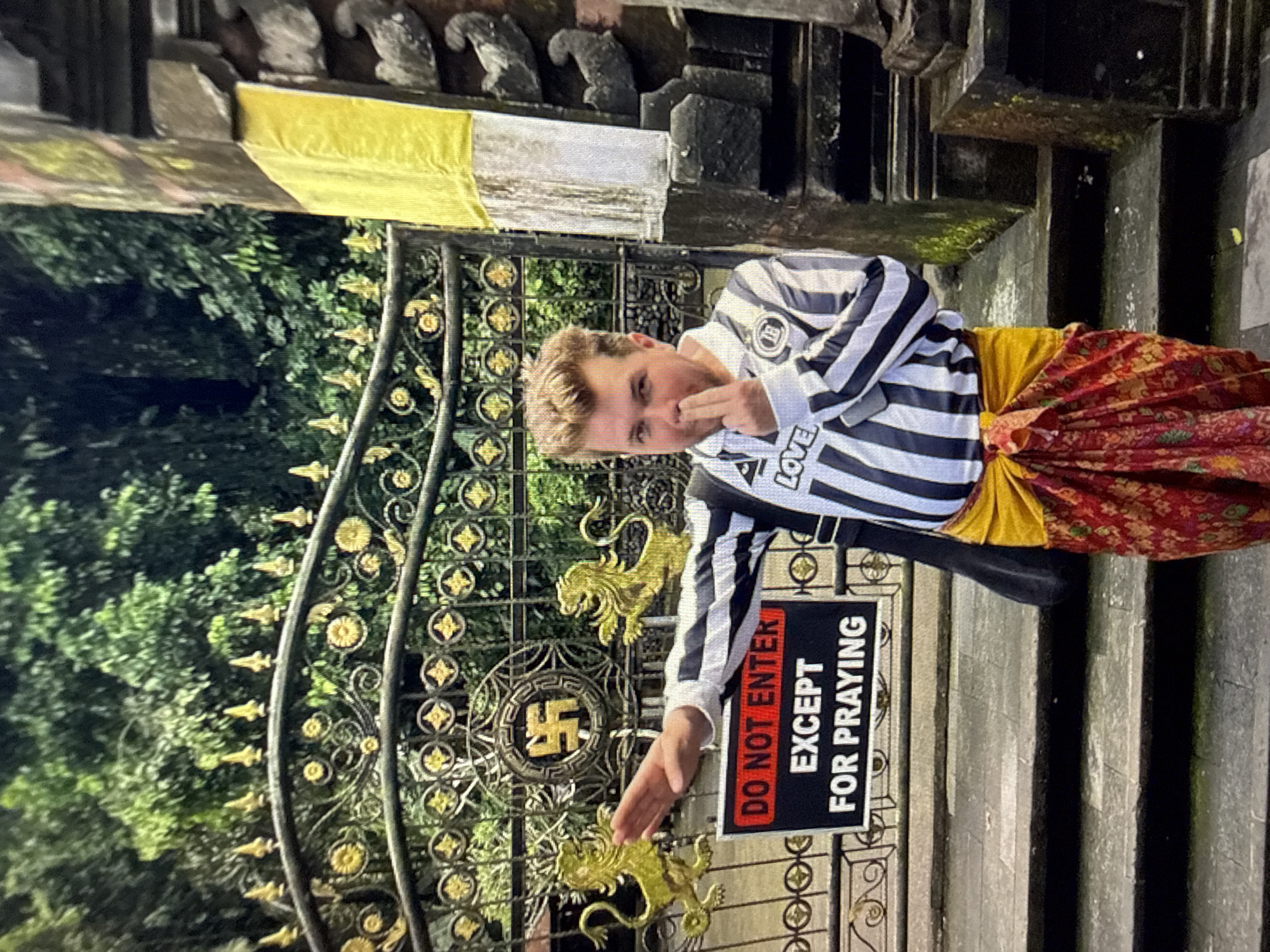The image depicts a Caucasian male standing in front of a gated entrance. The gate, made of wrought iron with gold-colored decorative elements, bears a symbol resembling a swastika in its design. A sign attached to the gate reads "DO NOT ENTER EXCEPT FOR PRAYING" in white text on a black background with a red bar. The man is dressed in a black and white striped shirt with the word "LOVE" on the lower part, a yellow sash around his waist, and a reddish-orange patterned cloth wrapped around his legs. The location is North Kuta, Indonesia. The gate opens onto a lush green area. Behind the gate is a stone structure with carvings. The image is rotated 90 degrees counterclockwise. The man has his finger under his nose.