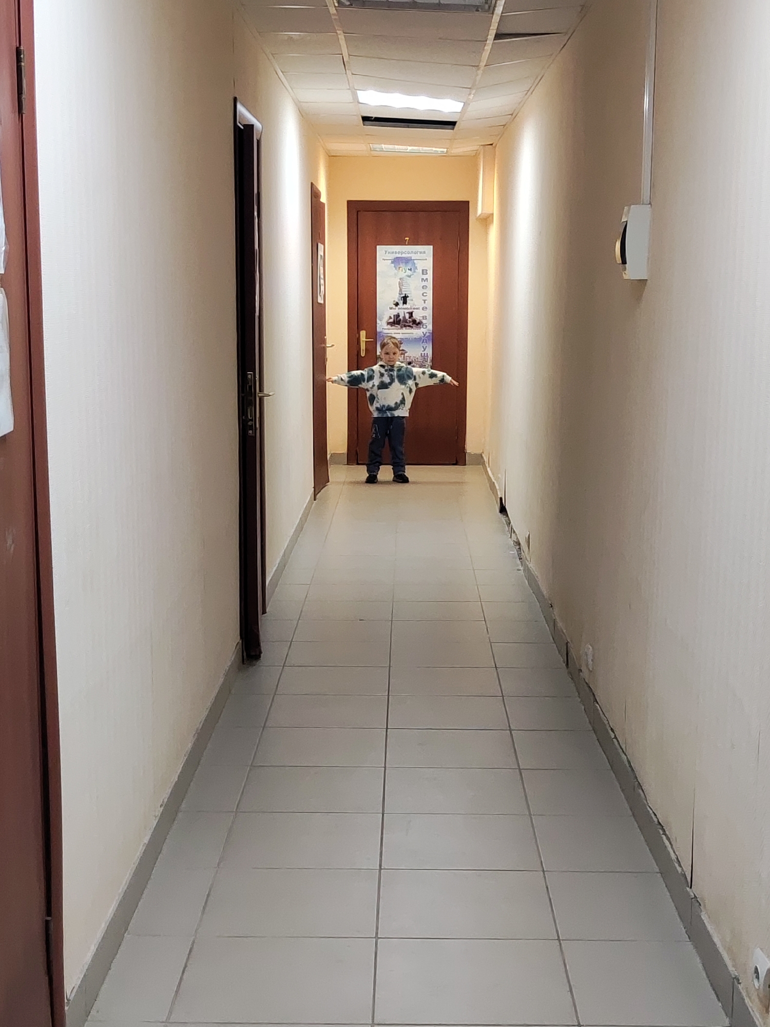 A young child stands centrally in a narrow, brightly lit indoor hallway. The child, approximately 4-6 years old, has light hair and is wearing a white, green, and blue tie-dye patterned long-sleeved top with dark pants and shoes. Their arms are outstretched horizontally, creating a "T" shape, suggesting a playful pose.
The hallway features light-colored (beige or off-white) walls with a dark grey baseboard running along the bottom. The flooring consists of light grey square tiles with darker grout lines. On the left side, two dark brown wooden doors are visible, with the closest one partially open. Another dark brown door closes off the end of the hallway, directly behind the child. A fourth door is partially visible on the right side in the background.
The ceiling is a suspended grid system with several panels appearing damaged or missing, revealing building infrastructure above. Fluorescent light fixtures are integrated into the ceiling, illuminating the space. On the right wall, a white rectangular electrical box with a vertical conduit is mounted, and further down, near the floor, a white electrical outlet is visible.
A poster is affixed to the door at the end of the hallway. Visible text on the poster, written in Cyrillic, includes "Универсалогия" (Universology) at the top, a large number "7", and phrases like "Мы создаем" (We create) along with a vertical list of words or acronyms. The setting appears to be a functional corridor in a building, possibly an office or institution in Chaikovsky, Russia, as indicated by the Cyrillic text and location context.