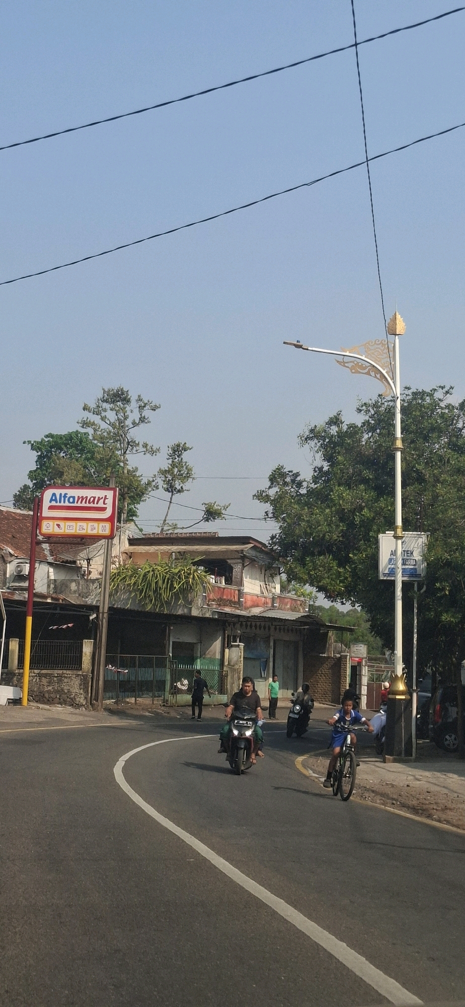 The image captures a street scene in Cilaku, Indonesia, under a clear blue sky. In the foreground, a paved road curves to the left, marked with a white line. Several people are depicted on various modes of transportation. A young boy on a bicycle rides on the right side of the road, heading towards the viewer. To his left, a man on a motorcycle is also moving towards the viewer, with a passenger behind him. Another motorcycle is visible further back on the left.
On the right side of the road, a decorative street light with an ornate golden element at the top stands tall. A sign attached to it reads "APOTEK" and "FARMA".
On the left side of the image, an Alfamart store is prominently visible, indicated by its red and white signage. A red and yellow pole supports the sign. Adjacent to the Alfamart, buildings with multiple stories are present, some with balconies and visible greenery. A chain-link fence encloses an area to the left of the buildings. Trees and vegetation are scattered throughout the scene, with a large tree dominating the background on the right. Power lines are visible crisscrossing the sky.