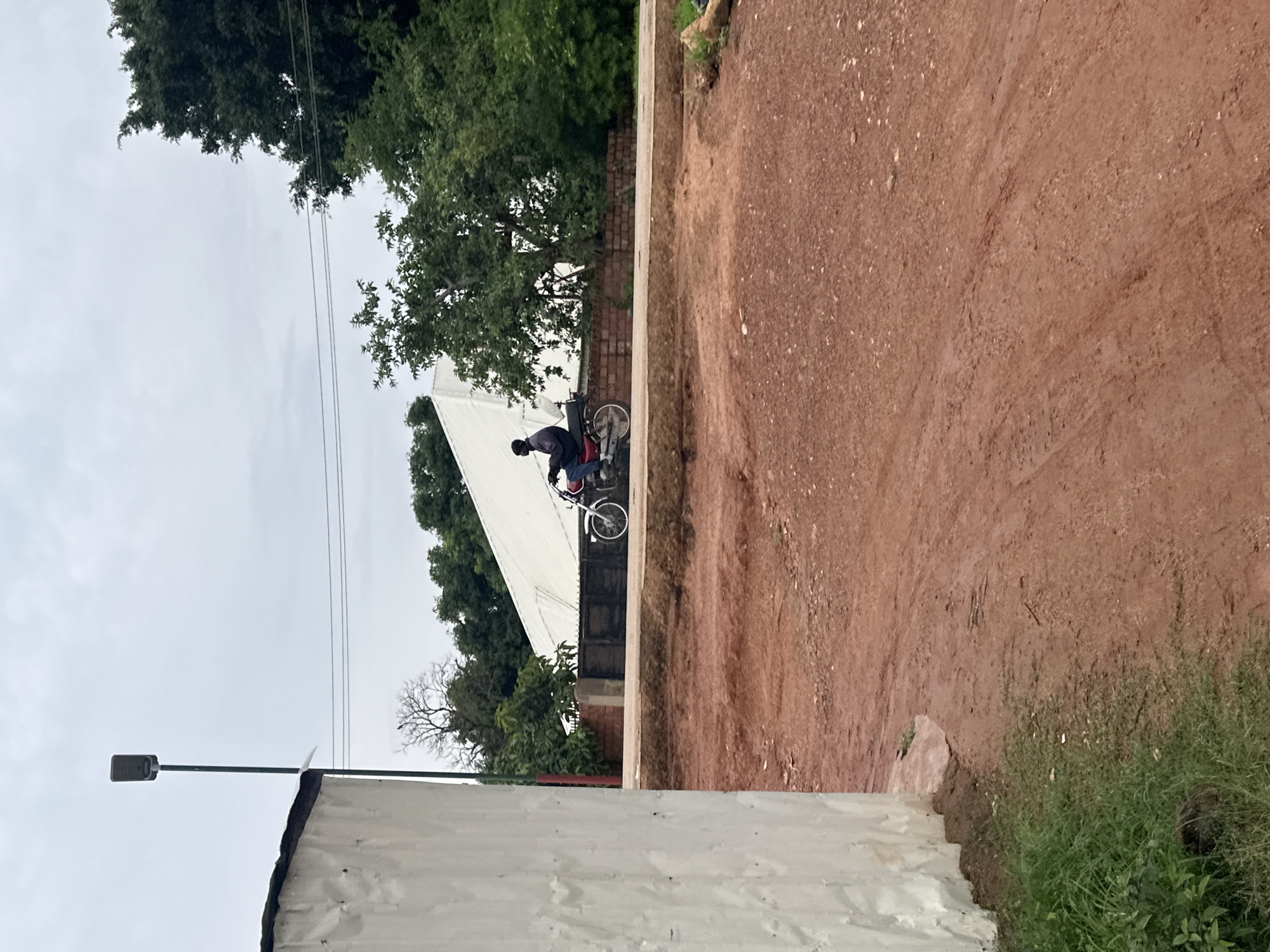 An overcast day in Du, Nigeria, depicts a person riding a red motorcycle on a paved road. The rider, wearing a dark jacket and blue pants, is captured mid-motion. To the left of the scene, a large, textured white wall dominates the foreground, partially obscuring a street lamp. Behind the motorcycle, a metal fence runs along a low brick wall, leading to a building with a white, angular roof. Lush green trees flank the background, creating a natural contrast with the man-made structures. The ground in the foreground is a rich, reddish-brown earth, suggesting a rural or semi-urban environment. The sky is a uniform, muted grey.