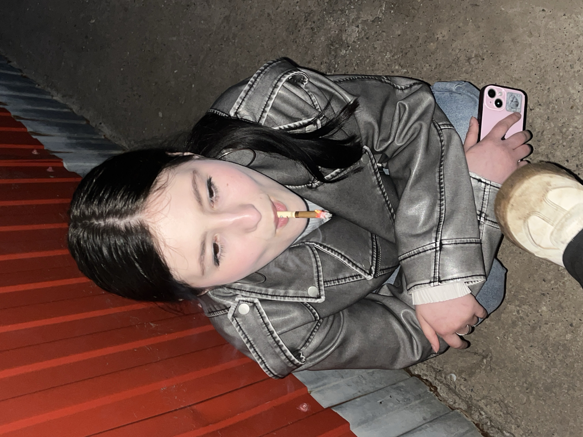 A young woman with dark hair is lying on the ground next to a red and grey corrugated metal wall. She is wearing a silver jacket, jeans, and a white top. She is holding a pink smartphone in her right hand and has a lit cigarette in her mouth. A white shoe is visible near her right hand. The location is not specified.