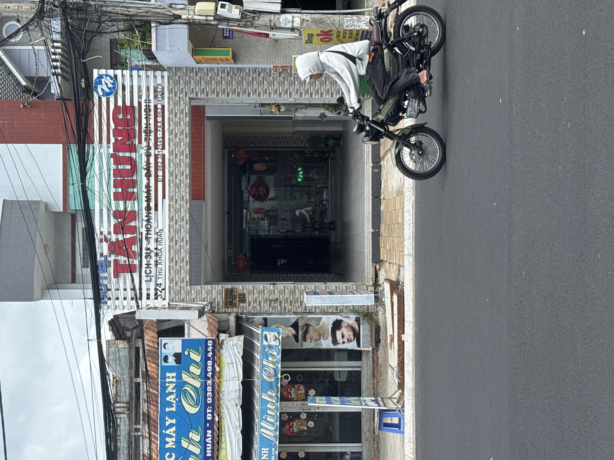 A person rides a motorcycle down a street in Phu Thuy, Vietnam. The person is wearing a grey hoodie and dark pants. The motorcycle is green and has wire-spoke wheels. The street is paved and has some debris on the side. Buildings line the street, including a shop with a sign that says "Minh Chi." There are also advertisements for haircuts displayed on the side of the building. Power lines are visible overhead. The sky is overcast.