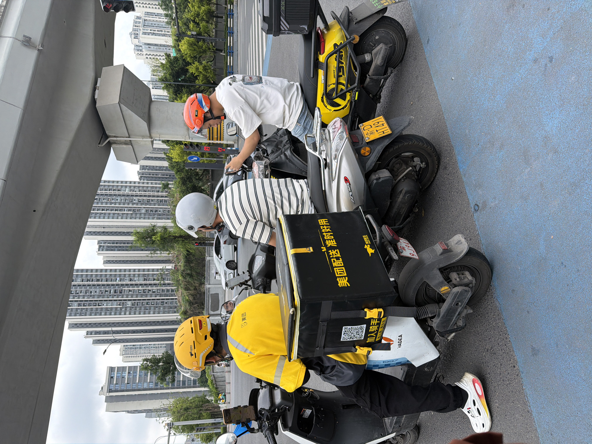 Three individuals on electric motor scooters appear to be engaged in a discussion near a road under a concrete overpass. The environment includes tall residential buildings, street lights, road markings, and traffic signals.
One individual wears a yellow helmet and a yellow jacket with a delivery box bearing Chinese text, likely related to a food delivery service called "Meituan." Another person is wearing a white helmet and a striped shirt. The third person has an orange helmet, white shirt, and jeans. The license plate of a scooter is visible, and the scooter has the designation "HT491." The background includes multiple high-rise buildings, suggesting an urban location in Chongqing High-tech Zone, China.
