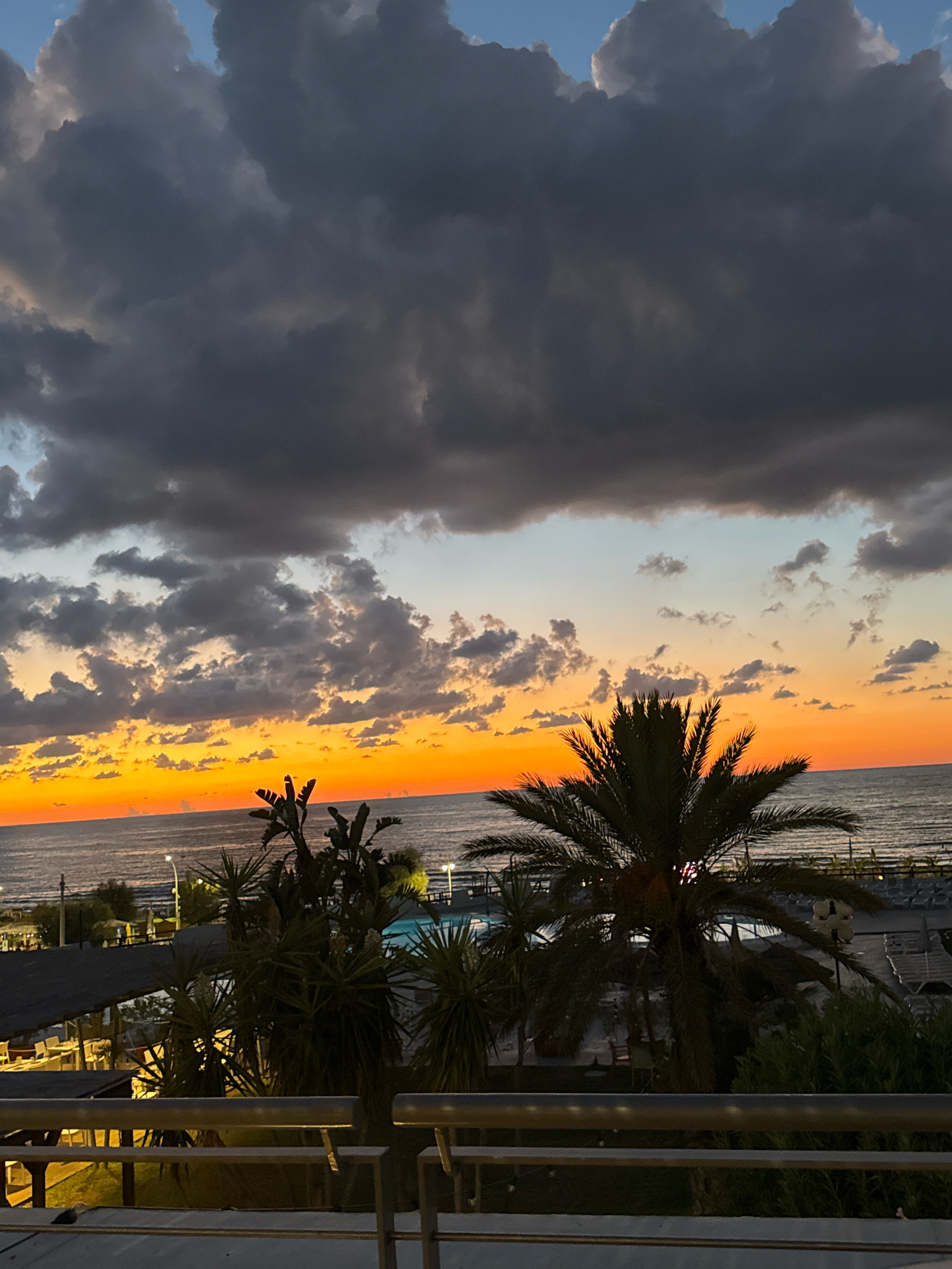 The image depicts a twilight scene over the sea at a resort location in Isola delle Femmine, Italy. The sky is dominated by dramatic, dark clouds that are beginning to break, revealing patches of bright orange and yellow sunset near the horizon. The sea is calm, with gentle waves reflecting the colorful sky. In the foreground, several palm trees and other lush vegetation are silhouetted against the vibrant sunset. A swimming pool is visible in the mid-ground, along with lounge chairs and part of a building with an awning, suggesting a resort or hotel setting. The overall impression is one of a beautiful and serene evening by the Mediterranean coast.