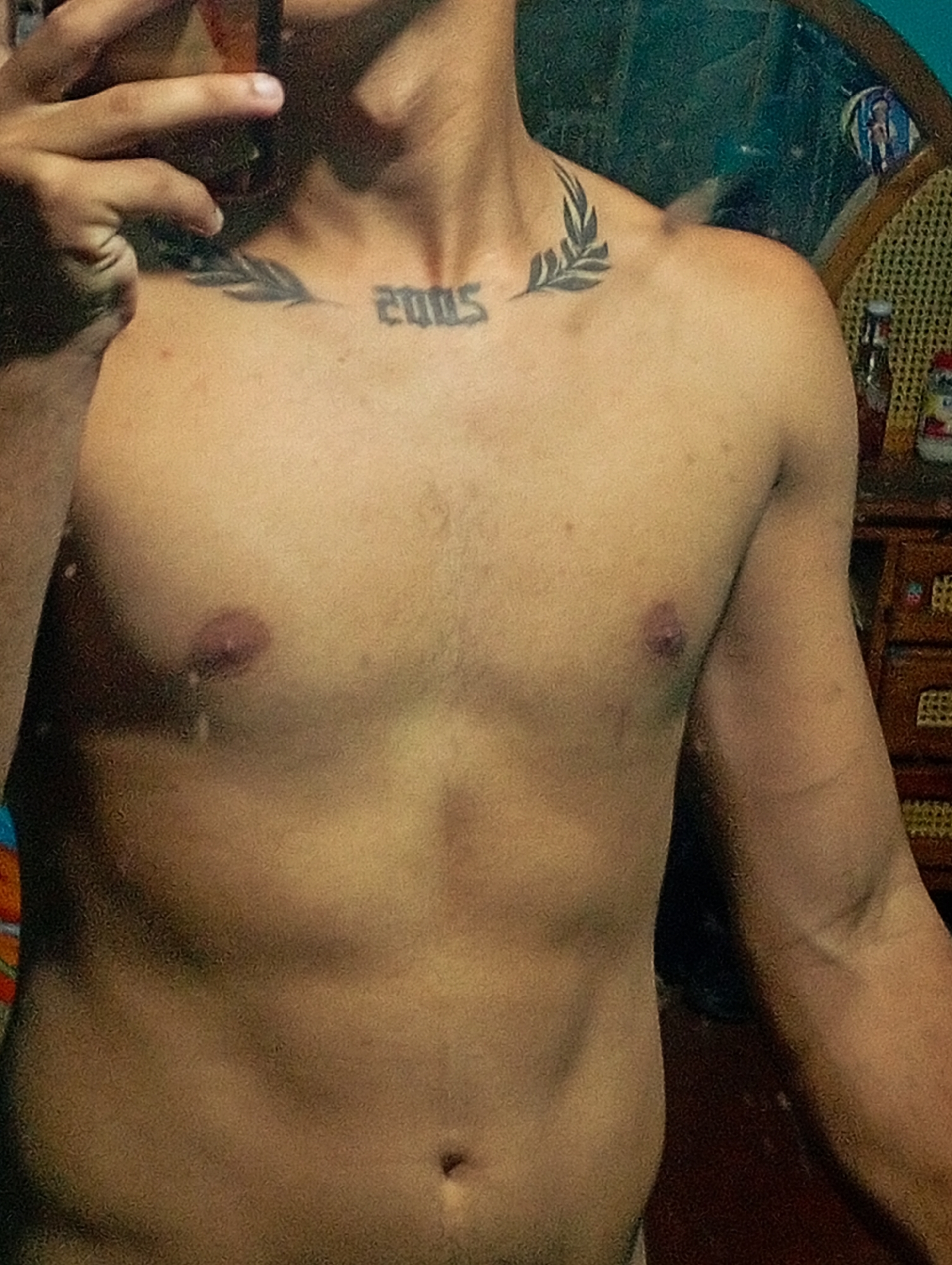 A shirtless, light-skinned person is depicted from the torso up. The person has a tattoo of the year "2005" in Gothic-style font between two leaf designs on their upper chest. A hand holding a phone is visible in the upper left corner. The background includes a mirror reflecting an interior scene with a dresser and various items. The location is in Altagracia, Venezuela.