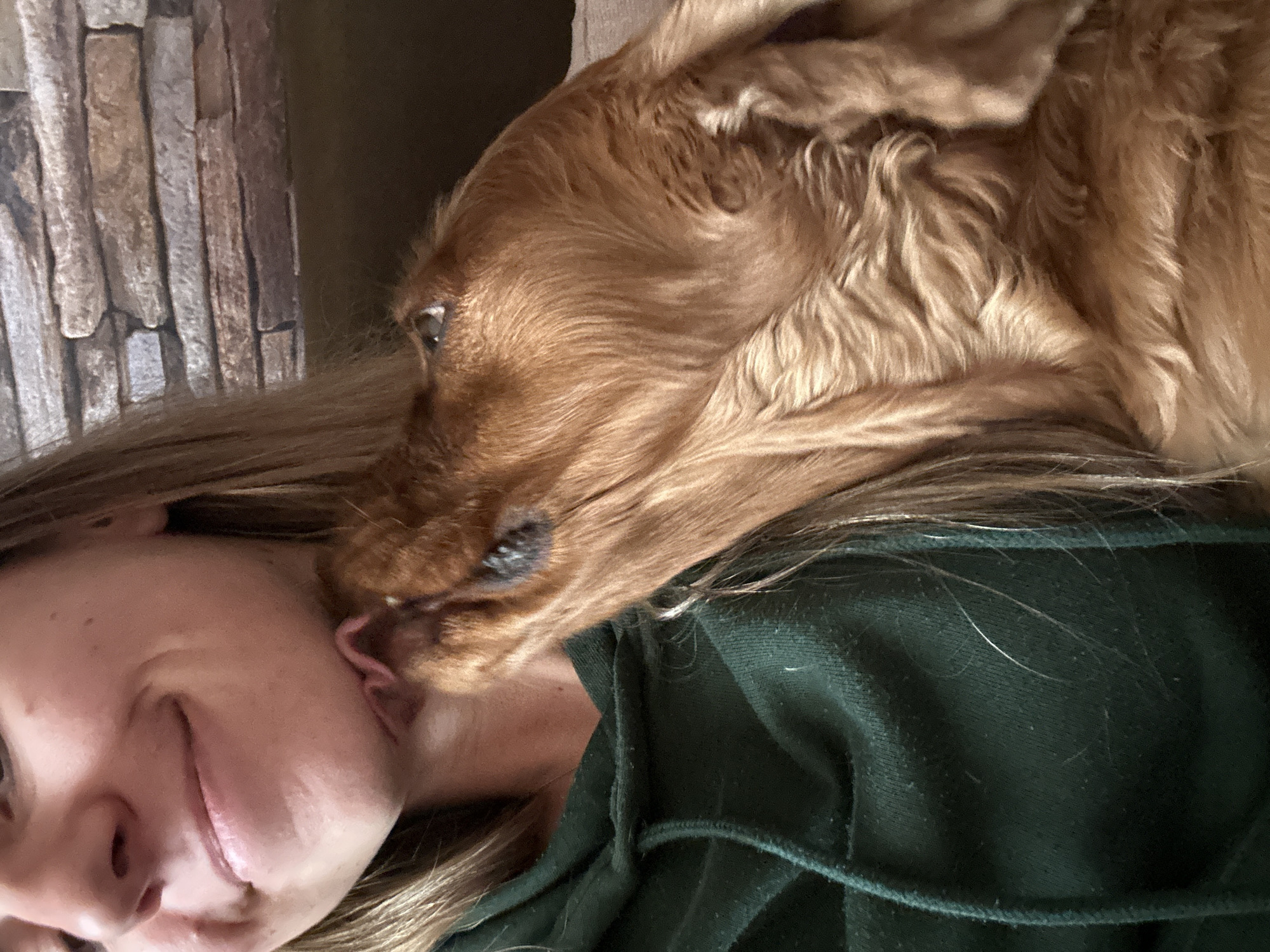 A person and a dog are interacting indoors. The person, a fair-skinned individual with blonde hair, is smiling. The dog, a golden cocker spaniel, is licking the person's ear. The person is wearing a dark green hoodie. The background includes a wall covered in what appears to be faux stone panels. The specific location in Sofia, Bulgaria is undetermined.