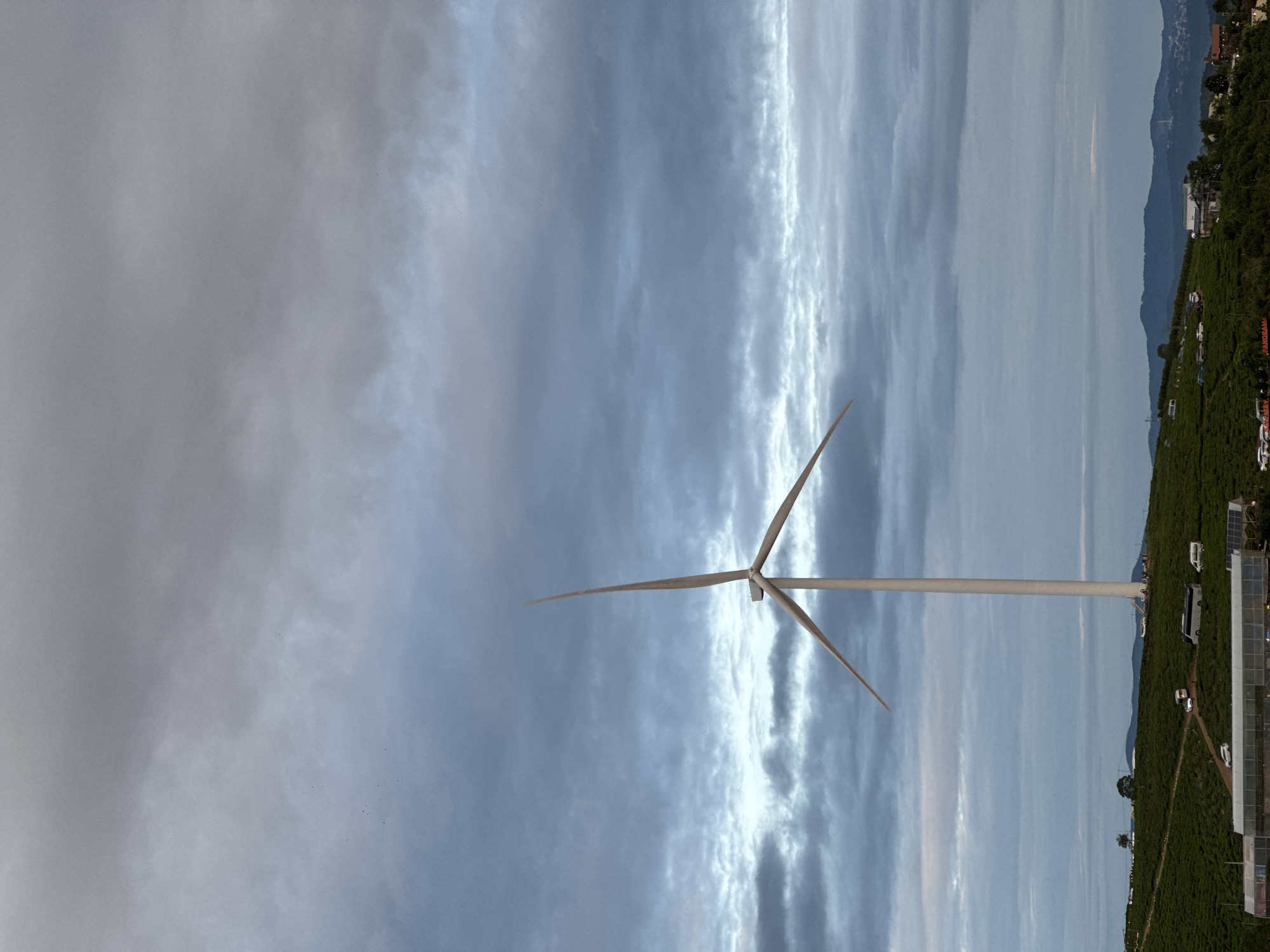 A vertically oriented photograph shows a wind turbine in Phát Chi, Vietnam. The turbine has three blades and is set against a striated cloudy sky. To the right of the turbine, a hill covered in dense green vegetation extends along the frame. Several buildings, structures, and vehicles are visible amongst the greenery. The left side of the photograph is dominated by a cloudy sky.