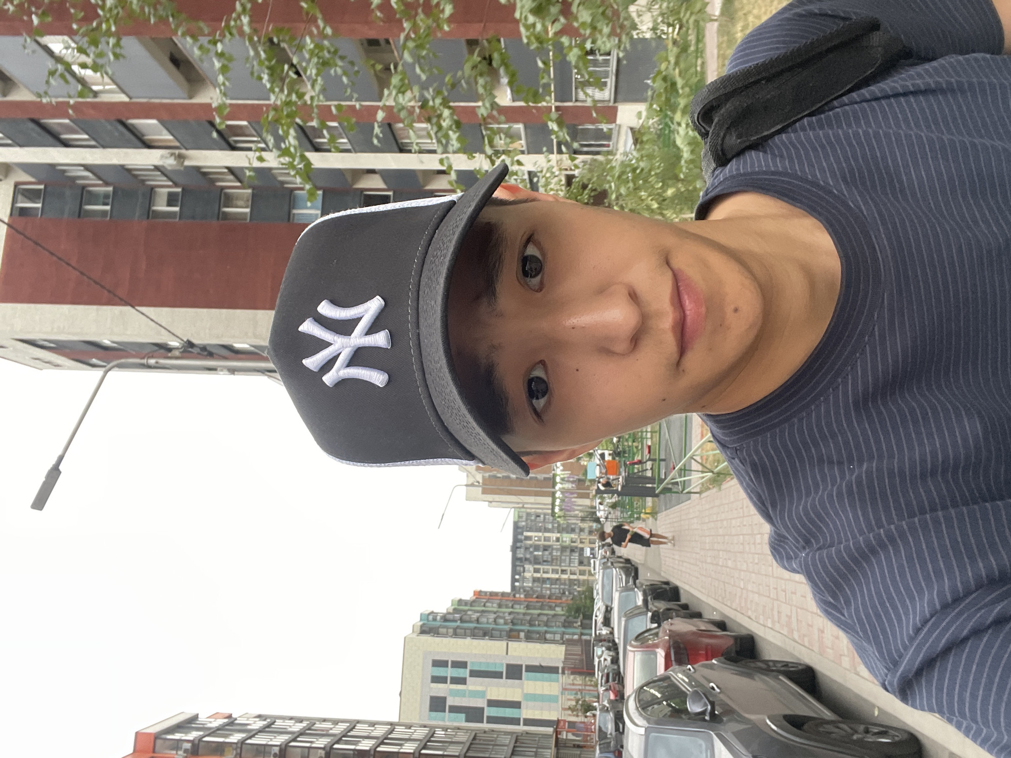 The image is a selfie taken by a young East Asian male wearing a black New York Yankees baseball cap and a dark blue striped t-shirt. He is standing on a paved sidewalk in a city, with apartment buildings and parked cars visible in the background. To his left, a row of cars is parked along the street. A lone male figure is walking away from the camera down the sidewalk. Behind the man, a children's playground with green fencing is visible. The buildings in the background appear to be modern, with red, gray, and multi-colored facades. The sky is overcast. The scene suggests an urban residential area in Almaty, Kazakhstan, characterized by its distinct architectural style of apartment blocks and well-maintained sidewalks.
