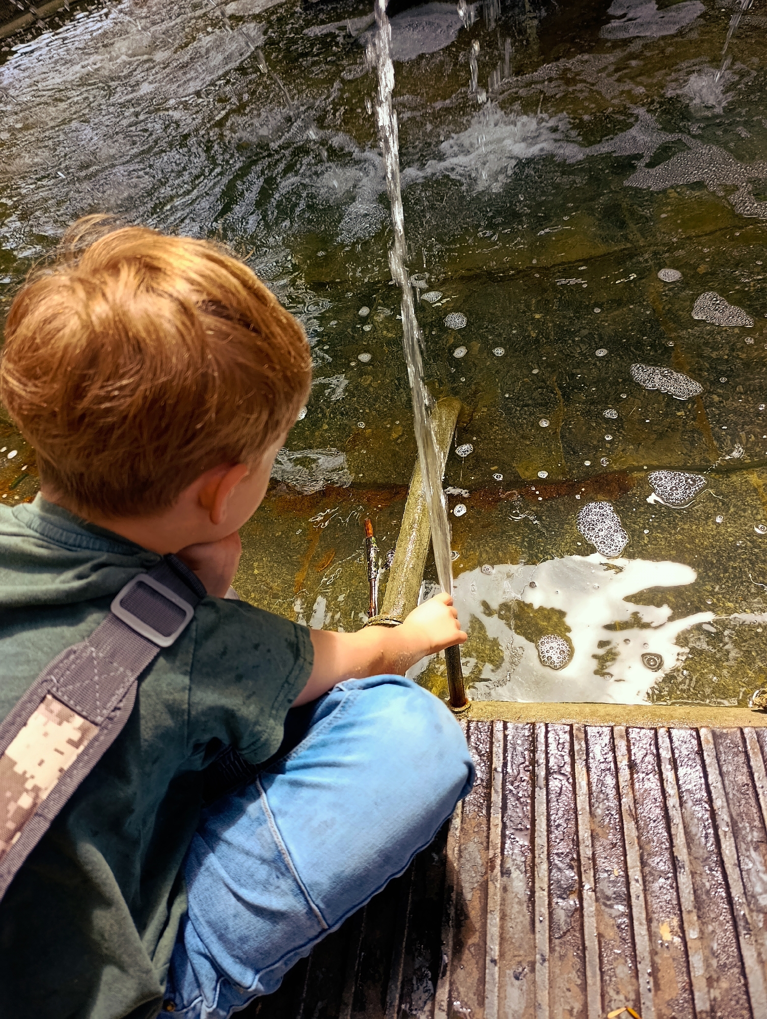 A young boy with auburn hair, wearing a green t-shirt and blue jeans, sits on a ribbed metal platform next to a shallow body of water. He is looking towards the water, with his right hand holding a wooden stick that is partially submerged in the water. A stream of water is pouring from above into the water. The water appears murky and has some bubbles and debris. The boy has a camouflage strap across his shoulder. The setting appears to be an outdoor area, possibly a park or a public fountain, but the specific location in Kyiv, Ukraine cannot be determined from the image.