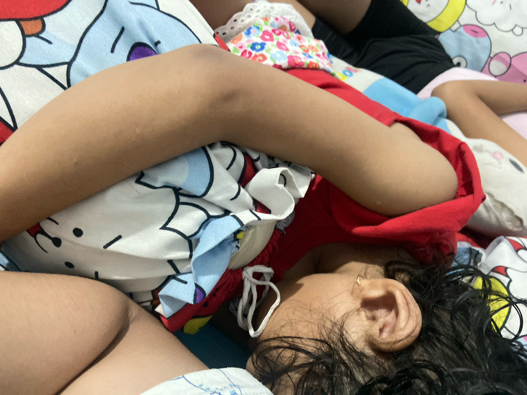 A close-up overhead view shows a child sleeping on a bed with colorful cartoon-themed bedding. The child is lying on their side, facing away from the camera, with their head resting on their arm. Their dark hair is spread across the pillow, and a small gold hoop earring is visible in their right ear. The child is wearing a red garment. The bedding features various cartoon characters, including what appear to be Hello Kitty and other animated figures in shades of blue, pink, yellow, and white. The scene is dimly lit, suggesting an indoor environment. No specific location in Gelam, Indonesia, can be determined from the image.