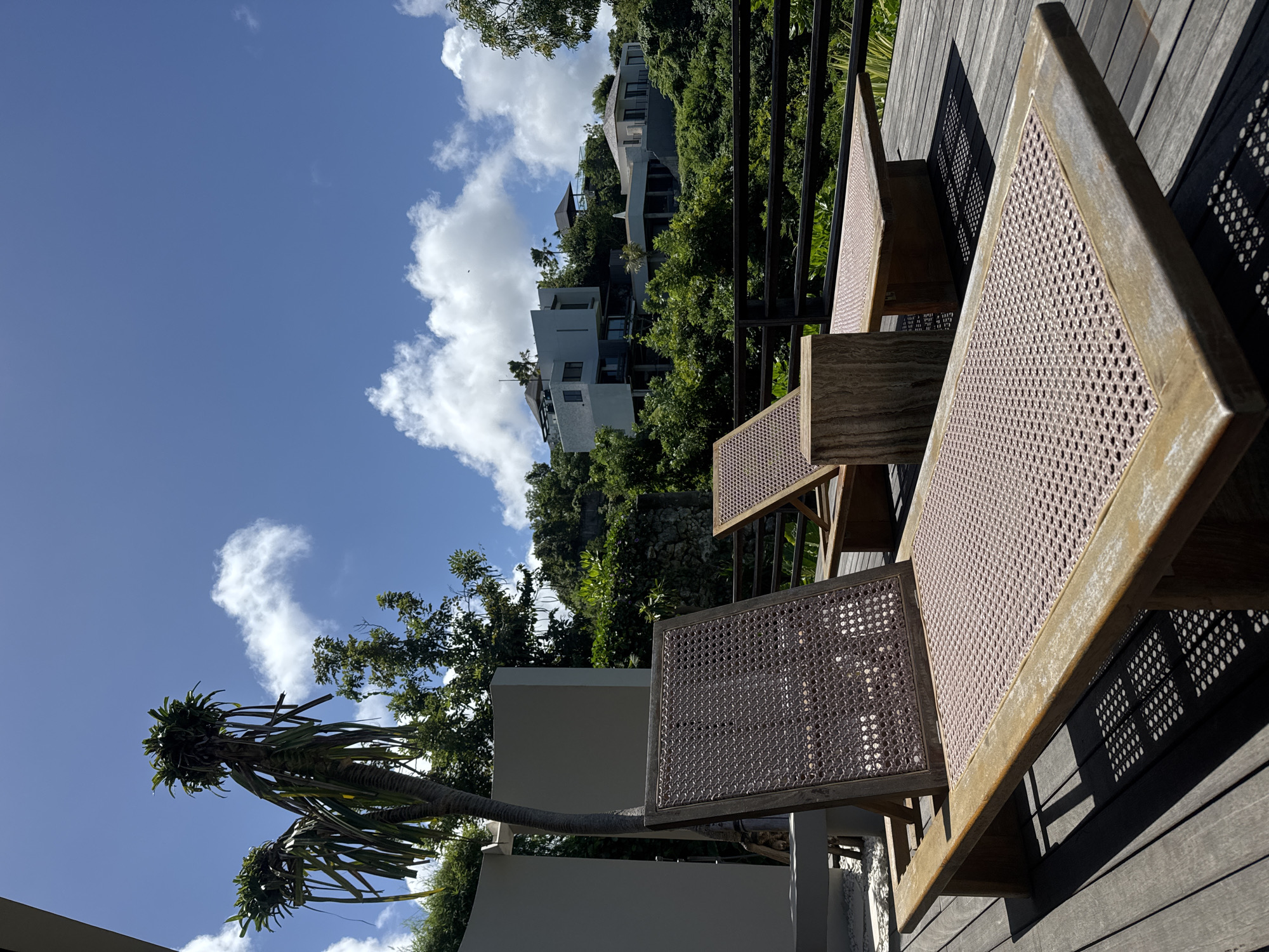 The scene shows outdoor furniture, specifically chairs with wicker-style seating, on a wooden deck in Kuta Selatan, Indonesia. The view includes a tropical sky with scattered clouds, lush green vegetation, and modern architecture built into a hillside. A palm tree is visible near a white wall in the foreground. Shadows cast by the furniture indicate bright sunlight. The setting appears to be a residential or resort area, showcasing a blend of modern design and tropical landscape.