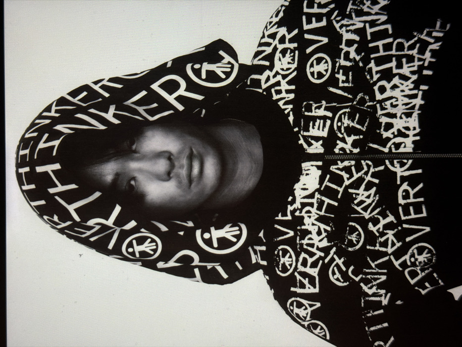A portrait-style photograph features a young adult male positioned against a plain, light-colored background. The individual exhibits East Asian phenotypic features, with dark hair parted centrally and falling to his chin, partially obscuring his forehead. His eyes are dark, and his facial expression is neutral. He is wearing a black hooded sweatshirt with the hood pulled up over his head. The garment, including the hood, is covered in a repeating pattern of white text and symbols. The visible text reads "OVER THINKER" in a distressed, block-lettered font. The symbols consist of a stylized human figure within a circle, often depicted with a large head or an indication of thought. A thin, metallic chain is partially visible around his neck beneath the sweatshirt collar. The image provides no specific environmental cues or background details to identify a particular location within the city of London, United Kingdom; it appears to have been taken in a controlled studio setting or against a neutral backdrop.
