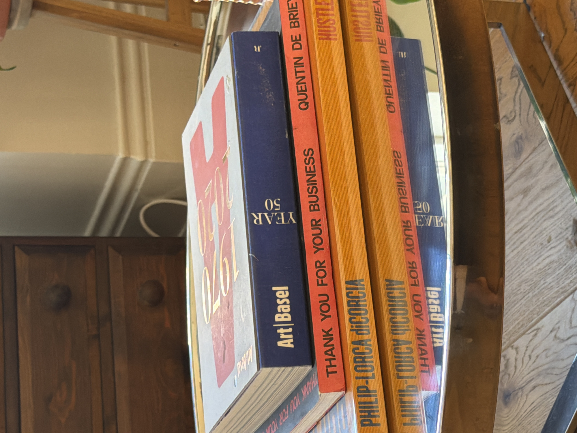 The image shows a stack of art books, with titles and authors visible on their spines. The books are placed on a reflective surface, which suggests they are perhaps in a room with mirrored elements. The visible titles include "Art Basel YEAR 50", "THANK YOU FOR YOUR BUSINESS", "PHILIP-LORCA DICORCIA", "HUSTLE", and "QUENTIN DE BRIE". The context of the location in Folkestone and Hythe District, United Kingdom, is not immediately apparent from the image. It could be in a private residence, an art gallery, or a bookstore, but this is not evident. The scene is focused solely on the stack of books and their immediate surroundings.