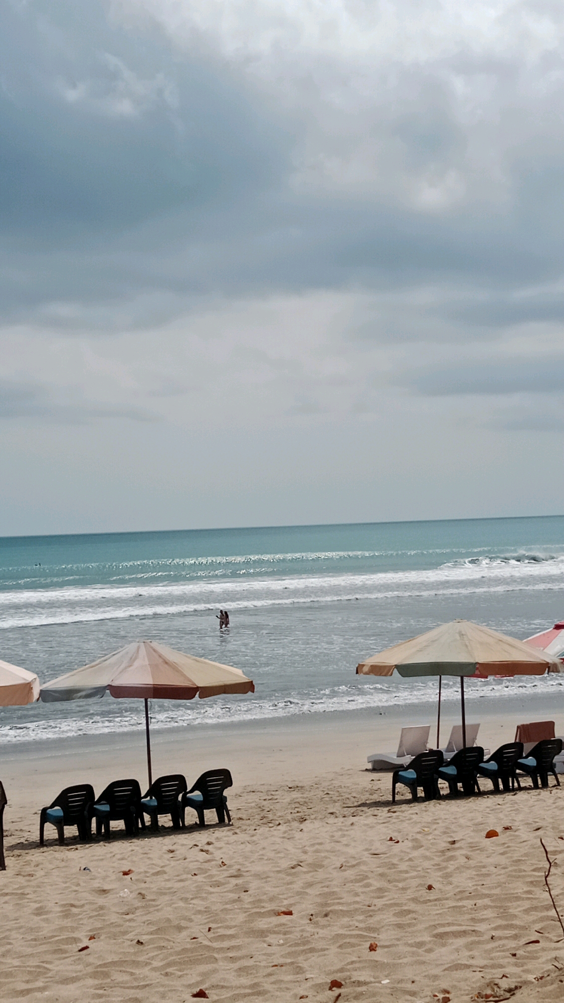 This image depicts a scene on Kuta Beach in Kuta, Indonesia, characterized by a cloudy sky and a calm ocean. In the foreground, the sandy beach is lined with beach chairs and umbrellas. Several black plastic beach chairs with blue cushions are arranged in groups, some under colorful umbrellas. Further back, white lounge chairs are visible. Two people are wading in the shallow water of the ocean. The sea exhibits gentle waves, and the horizon is marked by the meeting of the turquoise water and the overcast sky. The overall atmosphere is serene, suggesting a moderately busy day at the beach.