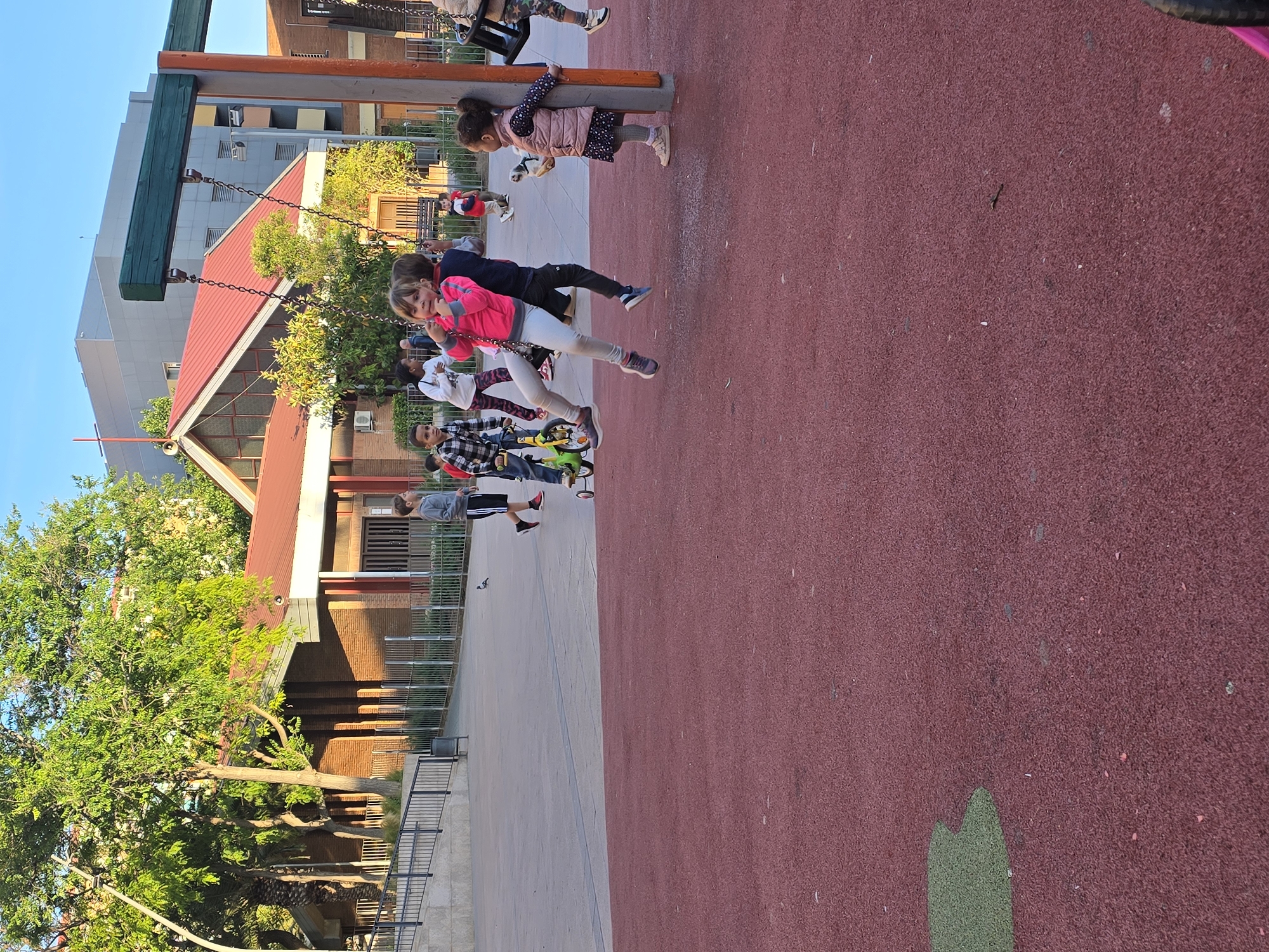The image depicts a playground in Cornellà De Llobregat, Spain. Multiple children are visible; some are on swings, and one is riding a small bicycle. The playground has a red surface. There are trees and buildings visible in the background.