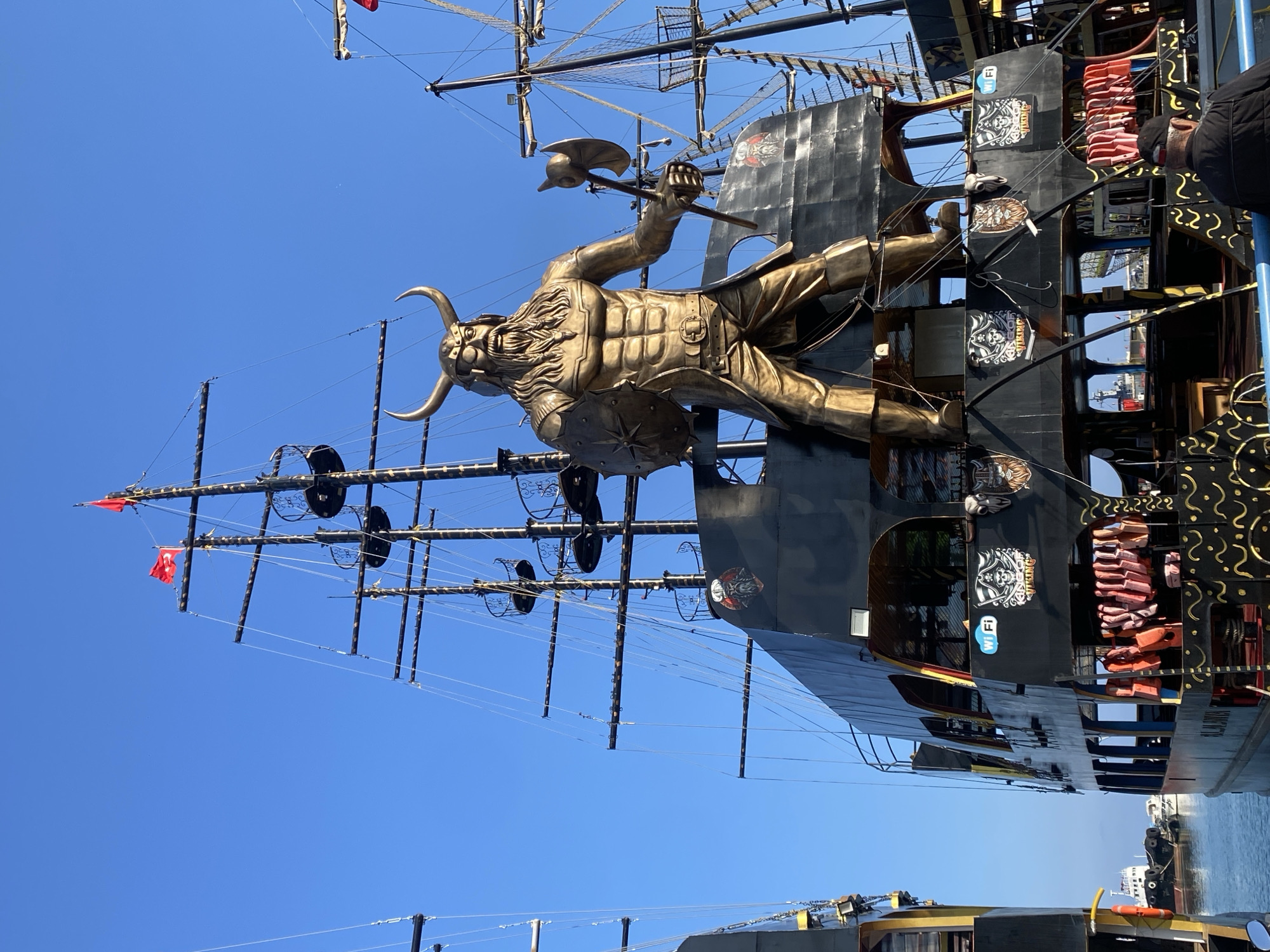 This media file captures a large, elaborately themed boat, resembling a Viking or pirate ship, likely moored in Alanya, Turkey. The central subject is a massive, golden-colored statue of a Viking warrior mounted on the vessel's side. The warrior figure is depicted with a horned helmet, a flowing beard, and muscular armor, wielding an axe in one hand and a shield with a star-like emblem in the other.
The boat itself features a dark (black or dark grey) hull with intricate golden, scroll-like decorative patterns, particularly noticeable towards the stern. It boasts multiple masts and complex rigging against a vivid, clear blue sky. On one of the upper decks, rows of neatly stacked orange life jackets are visible, indicating its use as a passenger vessel. A red Turkish flag, bearing a white star and crescent, can be seen flying from one of the masts, providing a clear geographical cue.
Visible text on the boat includes a "Wi Fi" symbol and an emblem featuring a skull with crossed weapons, below which the word "VIKING" is legible. The name "ALANYA" is also partially visible in gold lettering on the boat's stern, confirming the location context.
The scene is set on a bright, sunny day with a perfectly clear sky and calm, blue water. The water reflects the sunlight and portions of other boats or dock structures are visible in the background. The overall impression is one of a cheerful and adventurous tourist attraction, ready for a cruise. A person's head and shoulder are partly visible on an upper deck, possibly a crew member or passenger.