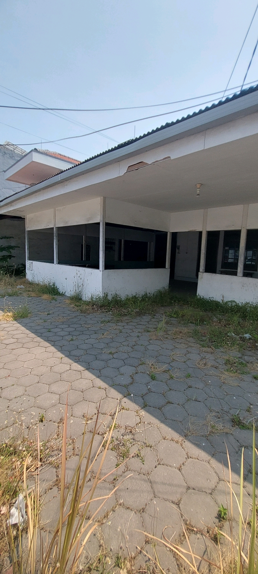 The image shows an abandoned, single-story building with a white exterior and a corrugated metal roof. The building has large, dark-tinted windows that appear to be boarded up or covered from the inside. The overhang of the roof shows signs of damage, with pieces missing. The foreground is a paved walkway made of hexagonal concrete blocks, with weeds and dry grass growing through the cracks. There are some tall, dry grass stalks in the immediate foreground. The sky is clear and blue. The environment appears to be a commercial or public space, likely in a state of disrepair. The location is identified as Surabaya, Indonesia. There are no people or active events depicted in the image.