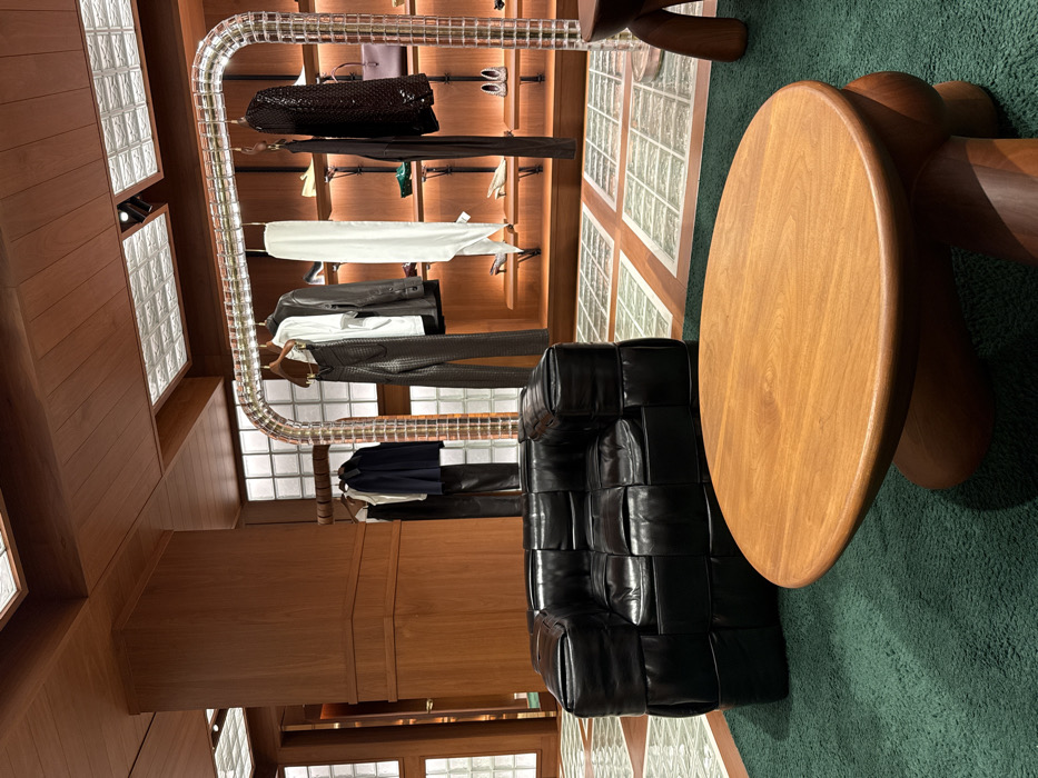 This image depicts the interior of a luxury retail boutique, identified as a Bottega Veneta store located at 12 Avenue Montaigne in Paris, France. The scene is devoid of people.
In the foreground, a low, circular wooden coffee table with a robust, sculptural wooden base rests on a dark green, plush carpet. To its left and slightly behind, a black leather armchair, characterized by a distinctive square-padded, modular design, is positioned.
The midground features a prominent, arch-shaped clothing rack constructed from clear, rectangular glass or acrylic blocks framed by metal. This rack displays various garments on wooden hangers, including a white t-shirt, dark leather trousers, a dark jacket, a white scarf, a black long-sleeved top, and a dark sequined jacket.
Behind the clothing rack, a wall-mounted display system with multiple illuminated wooden shelves showcases an assortment of luxury footwear, such as heels and flats, and handbags, including a dark brown purse. The store's interior is characterized by extensive warm-toned wooden paneling covering the walls and ceiling, including a large structural pillar on the left. The ceiling incorporates square recessed lighting fixtures with textured glass inserts. The floor beneath the carpeted area consists of a distinctive grid pattern alternating between wooden strips and translucent square glass blocks, which also appear in vertical panels along the back wall.