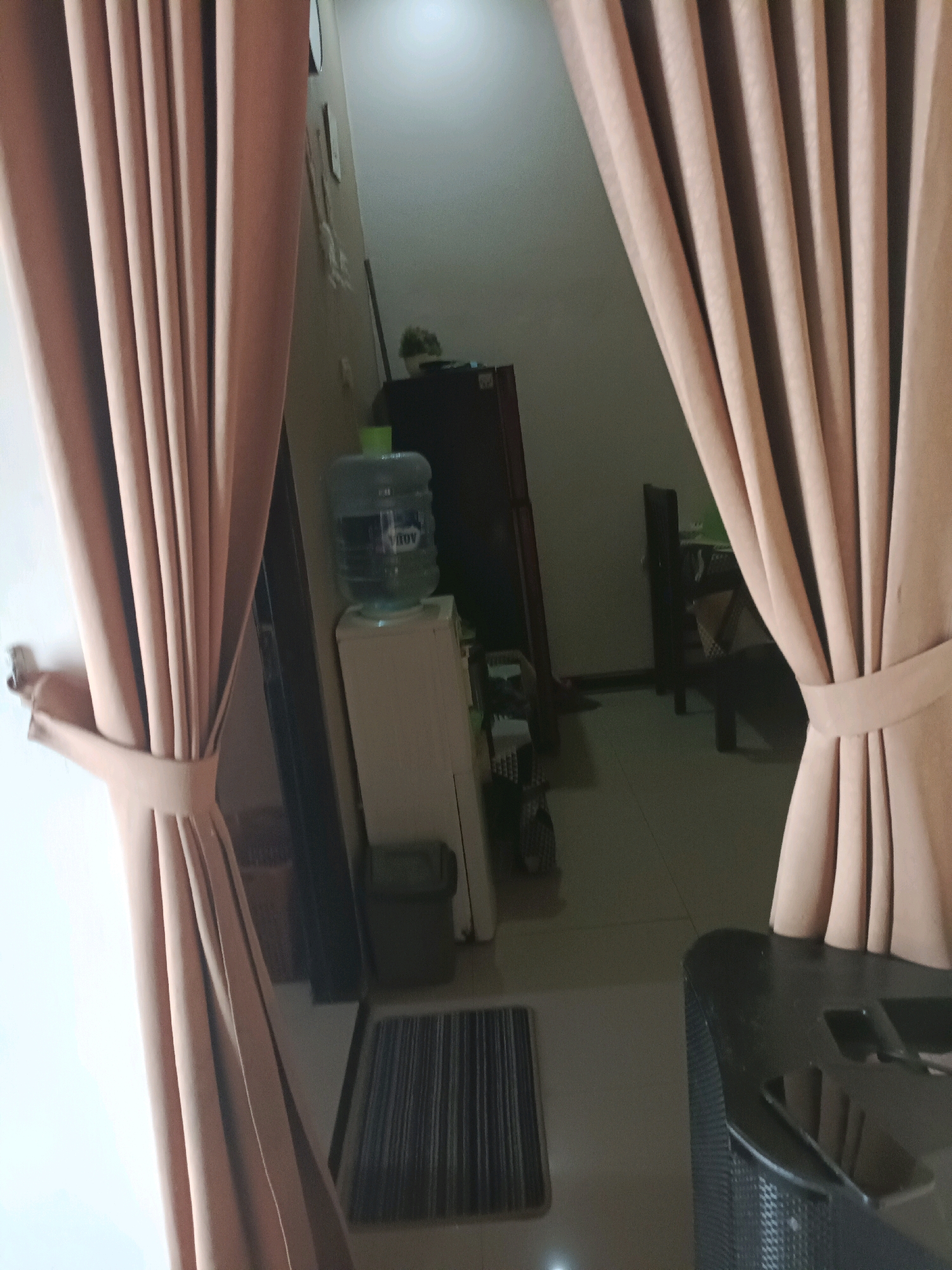 The image depicts an indoor scene, likely a room in a residence or an establishment in Rancang Kencono, Indonesia. In the foreground, two large, light brown curtains frame the view. The curtain on the left is partially drawn back, revealing a portion of a white wall. The curtain on the right is also drawn back, allowing a view into the background.
In the middle ground, a water dispenser with a large, clear plastic bottle filled with water sits atop a white cabinet. Behind the water dispenser, a dark-colored appliance, possibly a refrigerator, is visible. To the right of the water dispenser, a doorway or opening leads to another area.
The background shows a dimly lit space with what appears to be a table and chairs. The floor in this area is tiled. In the far right of the image, a dark, bulky object, likely a speaker or a piece of furniture, is partially visible, with several dark rectangular objects, possibly phones, resting on its surface. A ribbed doormat is situated on the floor between the cabinet and the dark object in the foreground. The lighting in the scene is generally dim, with some light emanating from above and to the left. There are no people visible in the image.