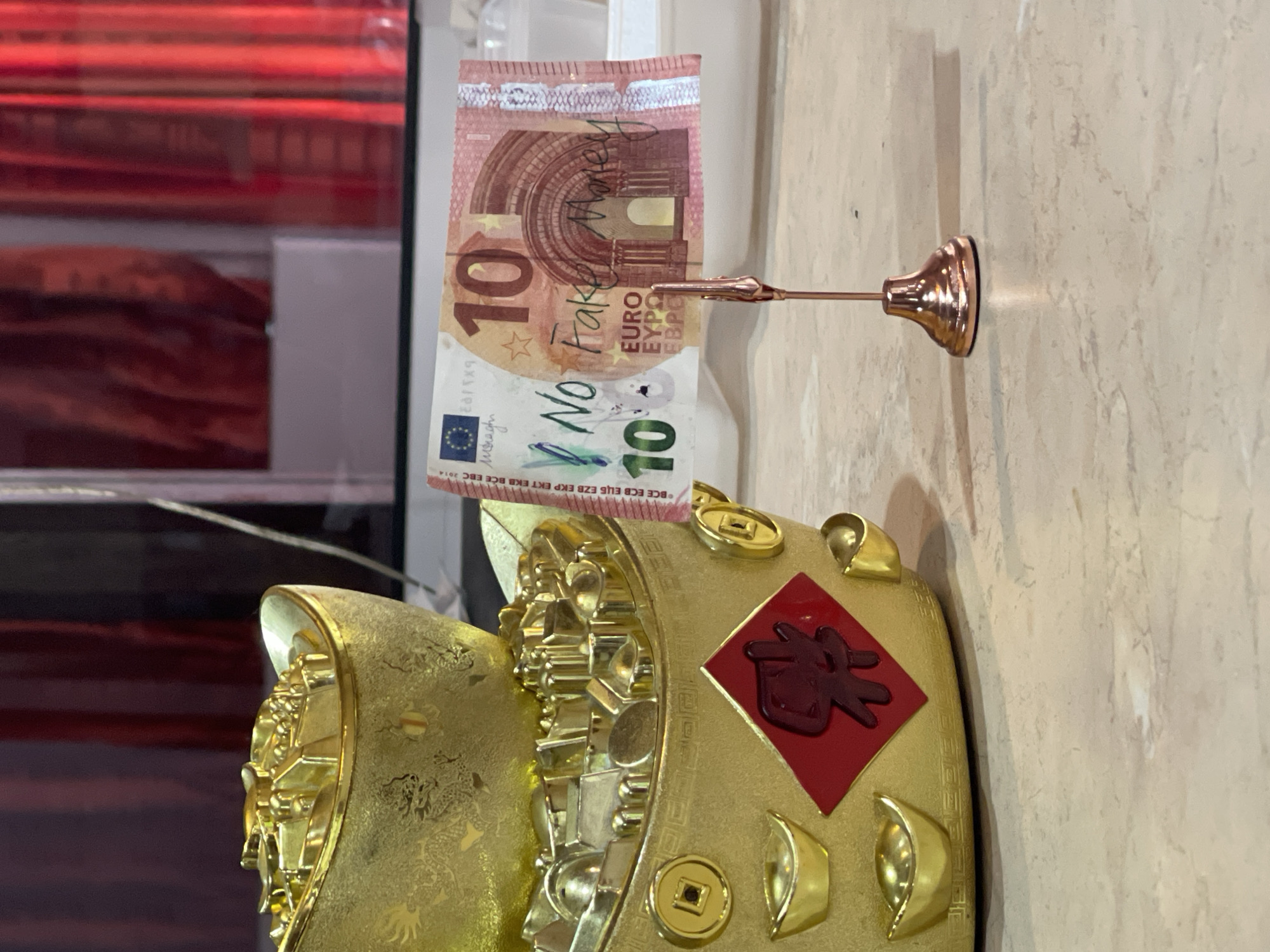 A close-up photograph shows a gold-colored figurine with Chinese-style decorations, including a red square with a character and small gold coin-shaped objects. A 10 Euro banknote with the word "Fake" written on it is held in place by a copper-colored clip attached to a stand. The background is blurred. The photograph was probably taken in Sofia, Bulgaria, as "Moragh" is Bulgarian currency.