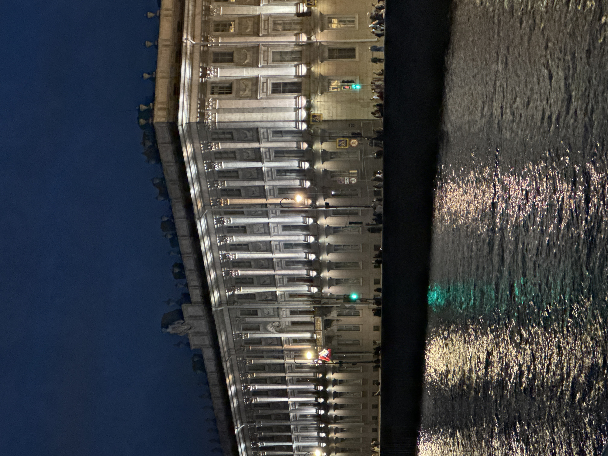 The image shows a grand building illuminated at night with a reflection in the water. The building is neoclassical in style, with a long facade featuring numerous columns and windows. It appears to be a public building or palace, possibly an art museum or government institution. The facade is brightly lit, highlighting the architectural details.
The water in the foreground is dark, reflecting the lights from the building and creating shimmering patterns on the surface. The sky above is a deep blue, indicating it is nighttime. There are some people visible on the embankment in front of the building, suggesting a public area. A flag is visible on a pole in front of the building. Traffic lights are also visible.
Based on the architectural style and the presence of the Neva River, this is likely the Winter Palace or a similar prominent building on the Palace Embankment in Saint Petersburg, Russia. The building's grandeur, the river, and the nighttime illumination are characteristic of this historic area.