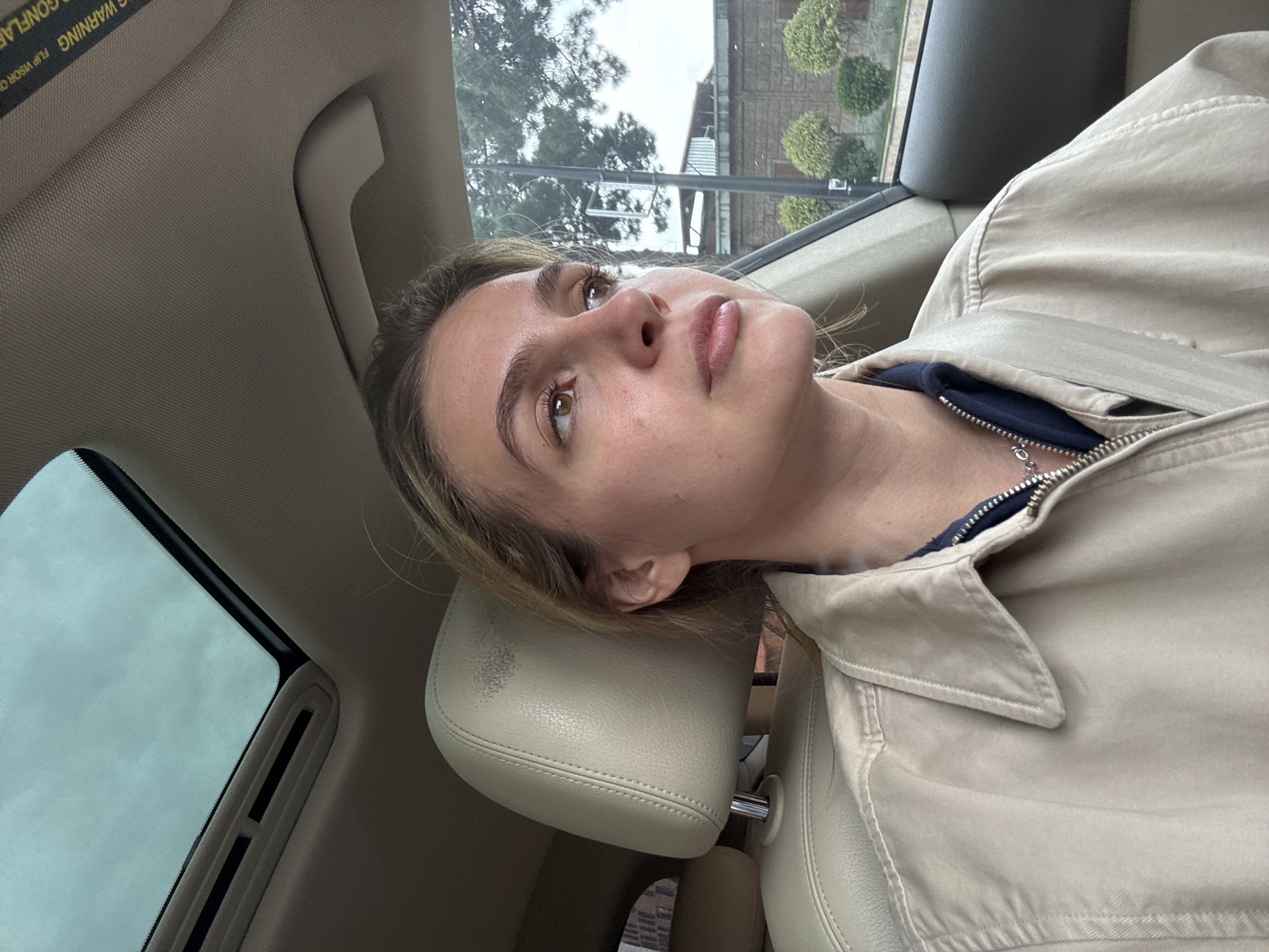 A young woman is depicted in a close-up, diagonally oriented shot, reclining inside a car. Her head rests against a light beige leather headrest, and her light brown hair is visible around her face. She has light brown eyes and full lips, with her gaze directed upwards and slightly to her right, conveying a thoughtful or relaxed expression. She is dressed in a light beige or khaki-colored jacket, partially unzipped to reveal a dark blue top and a silver necklace.
The car's interior features a light brown/tan headliner and trim. A sunroof is visible in the upper left corner of the frame, reflecting an overcast sky. A sun visor above her head shows partially visible, upside-down text that reads "WARNING. FLIP VISOR O". A grab handle is also present on the ceiling.
Through the car window, the exterior shows a cloudy sky, several trees with green foliage, and a building in the background, suggesting an urban or suburban environment. The overall lighting indicates a daytime setting under overcast conditions. The woman appears to be a passenger or relaxing within the vehicle.