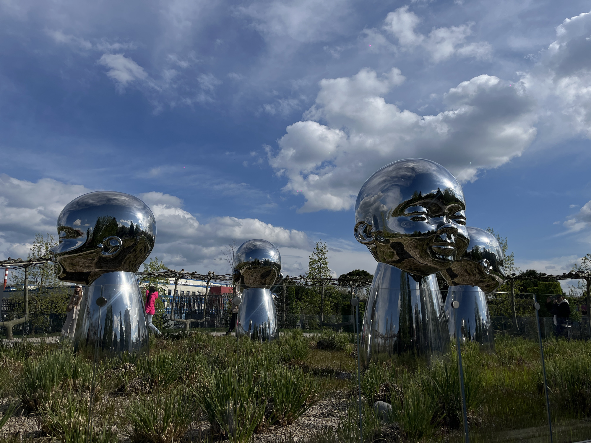 The image presents an outdoor art installation featuring several large, highly reflective metallic sculptures, each depicting a stylized baby's head atop a cylindrical body. These sculptures, resembling polished chrome, act as mirrors, reflecting the clear blue sky adorned with scattered white cumulus clouds, as well as the surrounding landscape and distant elements.
The sculptures are set within a cultivated outdoor space, characterized by patches of vibrant green, spiky ornamental grasses in the foreground and around the bases of the artworks. In the midground, trellises or pergolas are visible, along with various trees and shrubs. The background reveals architectural elements, possibly industrial buildings or urban infrastructure, under a partly cloudy sky.
A few blurry figures of people are discernible in the background, either walking through the area or captured as distorted reflections on the sculptures' shiny surfaces. One person in a bright pink top is seen walking behind a sculpture on the left, while another individual is faintly visible near a sculpture on the right.
The scene appears to be captured during the daytime, under bright natural light, suggesting pleasant weather with a mix of sun and clouds. The sculptures are monumental in scale, significantly larger than human height. Their highly polished surfaces create dynamic visual effects, merging the artworks with their environment through reflection. No discernible text is visible in the image. This scene is located in Krasnodar, Russia, likely within a public park showcasing contemporary art.