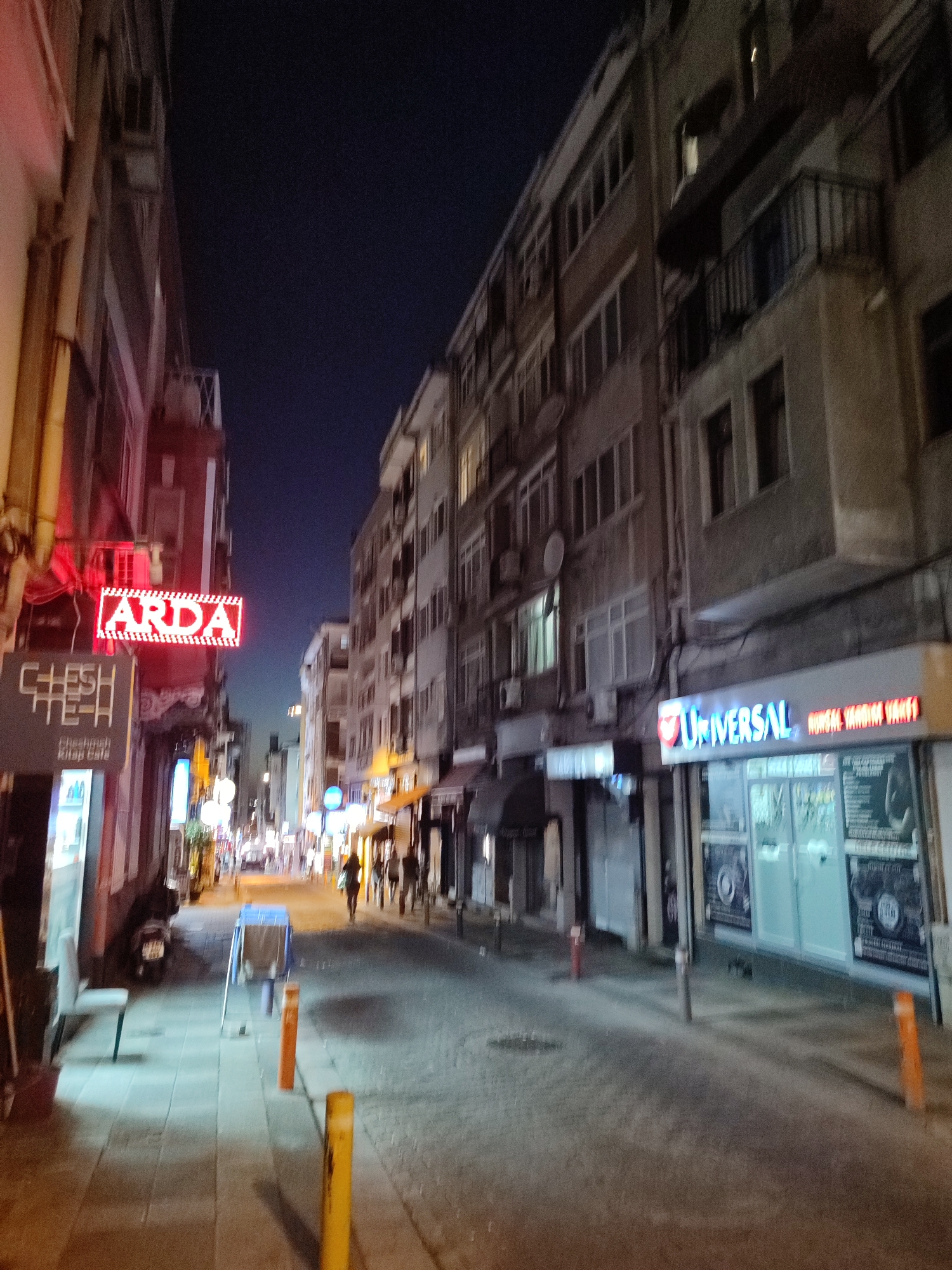 This is a night-time, ground-level, outdoor shot of a narrow street in Kadıköy, Turkey, likely a commercial or mixed-use area. The sky is dark blue, indicating it is late evening or night. Buildings with multiple floors line both sides of the street. The left side of the street features a building with a prominent red neon sign reading "ARDA" and a smaller sign below it that says "CHESHMEH Kitap Cafe." Further down the street on the left, a motorcycle is parked.
The right side of the street has businesses with illuminated signs and shop windows. One sign clearly reads "UNIVERSAL" in blue neon letters. Other businesses have posters or displays visible through their glass fronts. The street itself is paved with cobblestones in some areas and has a sidewalk. There are several bollards or short posts along the sidewalk.
Several people are visible walking on the street and sidewalk, appearing as silhouettes or indistinct figures due to the low light conditions. Some streetlights and building lights are on, illuminating the scene. The overall impression is of a bustling urban street at night.