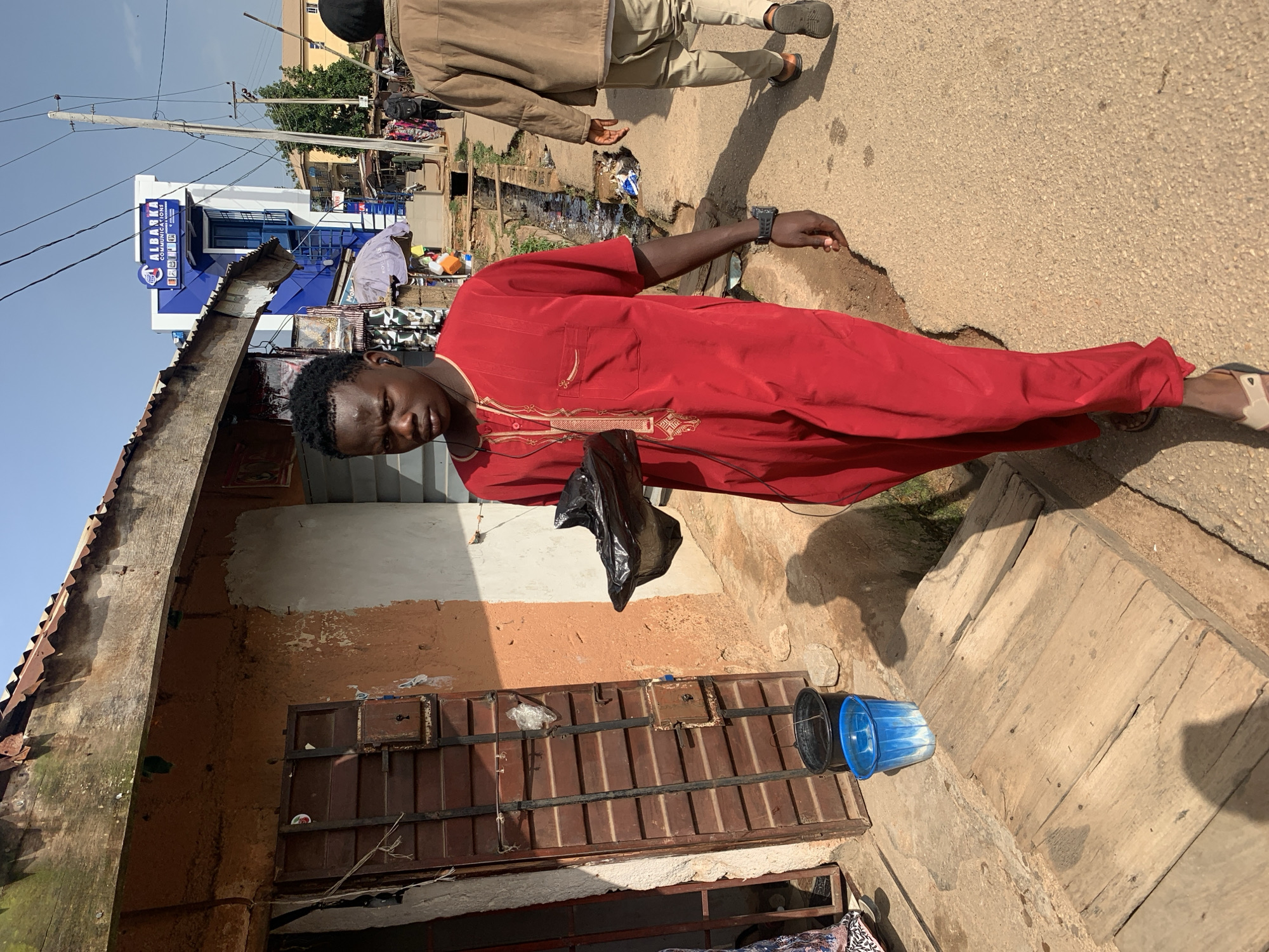 A young Black man wearing a red tunic and pants walks on a dirt path in the city of Gura Top, Nigeria. He is holding a black plastic bag in his right hand. A ditch filled with water runs along the right side of the path. In the background, a modern blue and white building with "ALBARKA COMMUNICATIONS" written on it stands out among smaller, more traditional structures. The sky is clear and blue. The overall scene depicts a typical urban environment in Nigeria.
