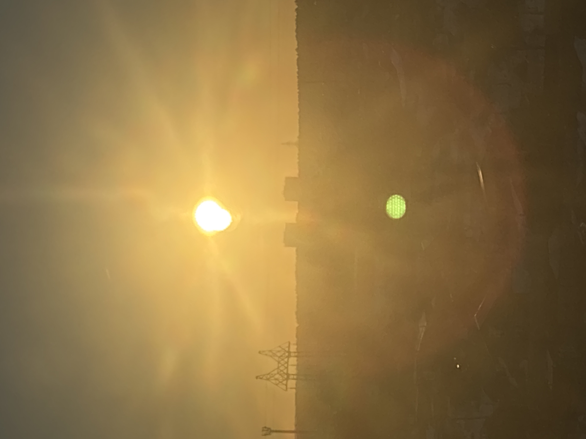 The image presents an urban and industrial landscape in Chaikovsky, Russia, illuminated by a low-angle sun. The sun, a brilliant white sphere with pronounced starburst rays and a large halo, is centrally positioned in the upper half of the frame. The sky is a uniform golden-orange hue, appearing hazy and diffused.
In the foreground, the lower portion of the image is dark and indistinct, featuring blurred, shadowed forms that suggest rooftops or industrial structures, partially obscured by atmospheric effects and lens flare. A prominent, circular, grid-patterned green lens flare is visible in the lower-left quadrant, and a smaller, bright white point flare is present near the center of the lower half.
Along the hazy horizon line, several major silhouettes are discernible. To the left, a tall, slender vertical pylon or tower stands. Further right, a large, distinctive steel truss structure, characteristic of a bridge segment (consistent with the Kama River Bridge), dominates the central-left horizon. To the right of this bridge-like structure, two rectangular, block-like building silhouettes are visible. Furthest to the right, a slender, conical structure, possibly a chimney or another tower, rises above the horizon. The overall scene is devoid of discernible people, actions, or specific interactions, with the environment heavily influenced by the intense light source and atmospheric haze.