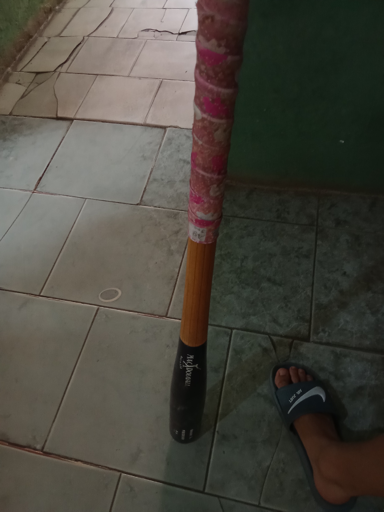 A baseball bat stands on a tiled floor next to a foot wearing a Nike slide sandal. The bat is dark at the bottom, transitioning to a wooden color towards the middle, and has pink and white tape wrapped around the handle. The floor tiles are grey and cracked in some areas. A green wall is visible in the background. The location is Juan Griego, Venezuela.
