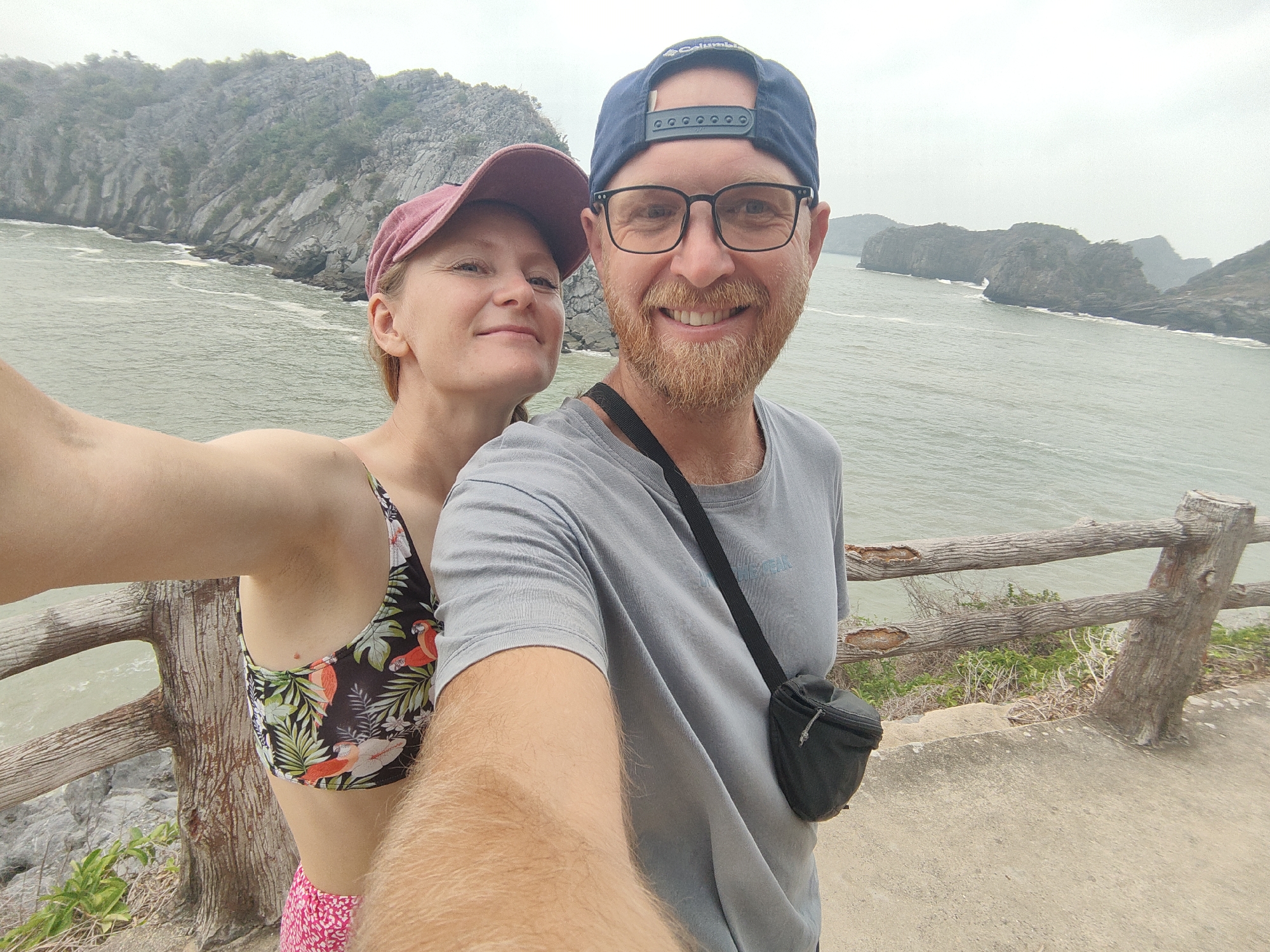 A man and a woman are taking a selfie on an overlook at Cát Bà Island, in Hải Phòng, Vietnam. The man, on the right, is wearing a blue baseball cap, glasses, and a gray shirt with a black fanny pack. He has a red beard and a broad smile. The woman, on the left, is wearing a pink baseball cap, a floral bikini top, and pink shorts. The backdrop features a body of water and rocky islands. A weathered wooden railing is visible in the foreground. The sky is overcast.