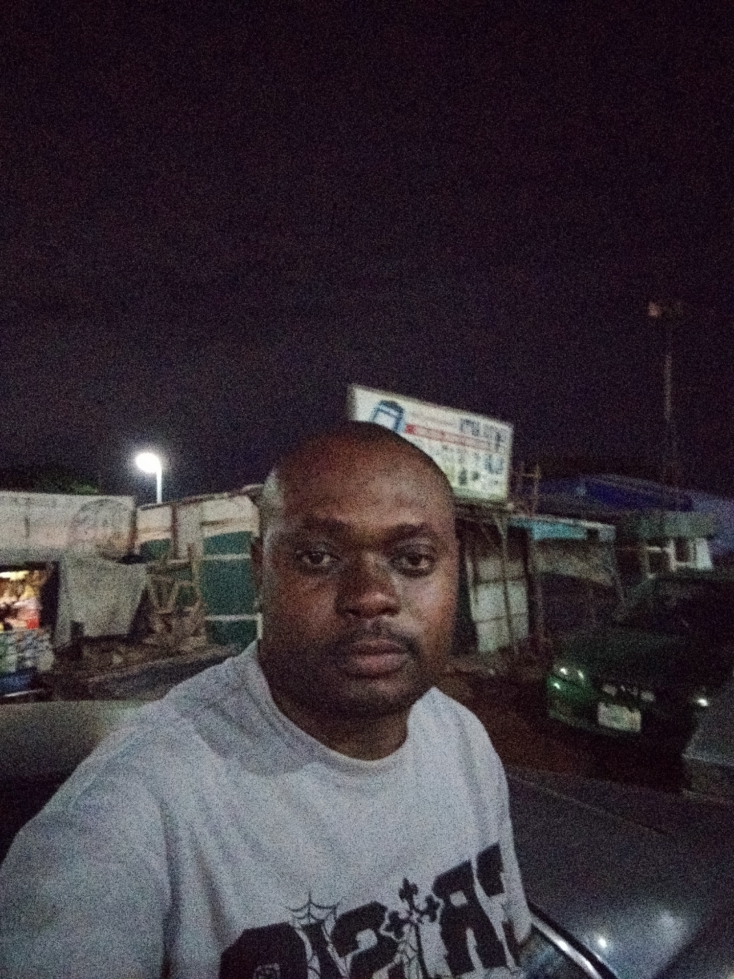An adult male of dark complexion, with a shaved head and a short beard, is positioned in the foreground, gazing directly at the camera. He wears a light grey or white short-sleeved t-shirt featuring a dark, partially visible graphic design on the chest. The scene is set at night in Pasere, Nigeria. In the background, a bright, singular street light illuminates several rudimentary, single-story structures or market stalls. Light emanates from within some of these structures. To the right, a large signboard with a blue and white logo or text is affixed above additional background buildings. A dark-colored vehicle, possibly a sedan, is parked on the right side of the street, with its rear facing the viewer. The entire image exhibits significant digital noise, indicative of low-light conditions.