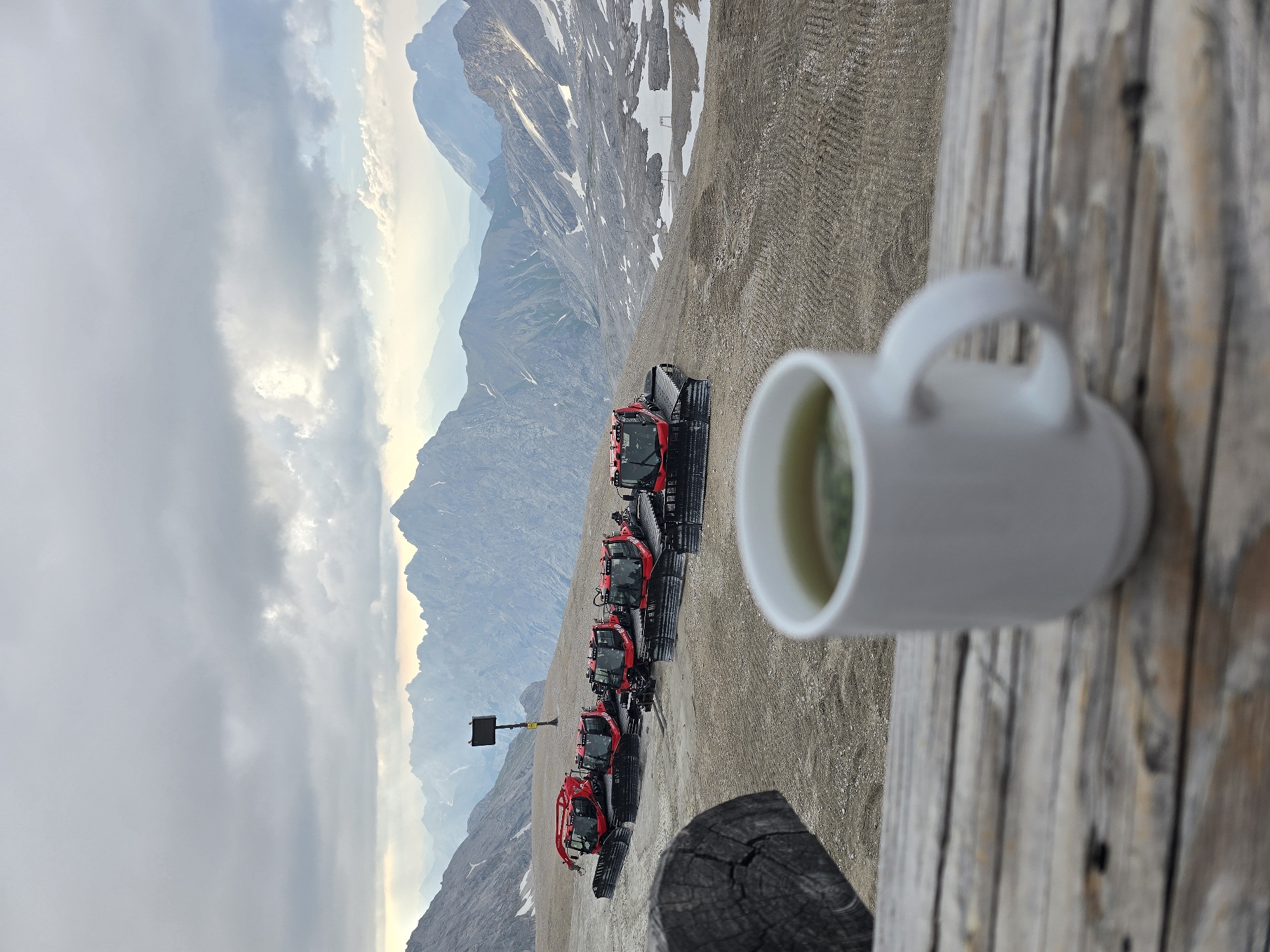 The image shows a scenic mountain landscape in Garmisch-Partenkirchen, Germany, with a line of red snow groomers parked on a gravelly slope. In the foreground, a white mug filled with a light-colored beverage, possibly tea, is placed on a weathered wooden surface, partially obscuring the view of the snow groomers. The mountains in the background are majestic, with some patches of snow still visible on their peaks. The sky is partly cloudy, with soft sunlight filtering through, suggesting it might be early morning or late afternoon. The overall atmosphere is serene and peaceful, with a sense of tranquility and breathtaking natural beauty. There is no visible text in the image.