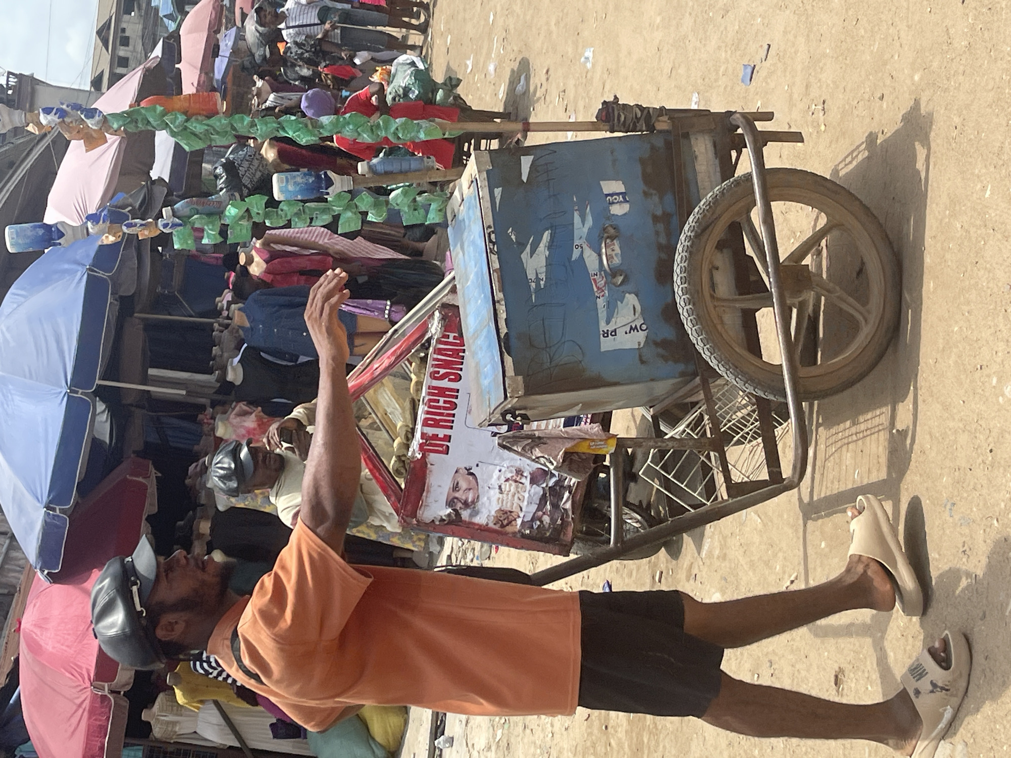 A ground-level, full shot depicts a man in an orange shirt and black shorts standing next to a food cart. The location appears to be a bustling market or bazaar in the city of Unknown, Unknown.
The man wears a dark cap and tan sandals. He faces left, with his left arm raised. The food cart is to his right. The cart, constructed of metal bars, has a large single wheel. A blue container sits atop the cart, covered with partially peeled stickers and posters. A sign reading "DE RICH SNACK" is attached to the front of the cart.
The background shows a dense crowd of people and market stalls with various items for sale, including clothing and umbrellas in shades of blue, pink, and red. Green and clear plastic bottles hang above the stalls. The ground is an uneven tan color, possibly dirt or sand. The overall lighting suggests a bright, sunny day.