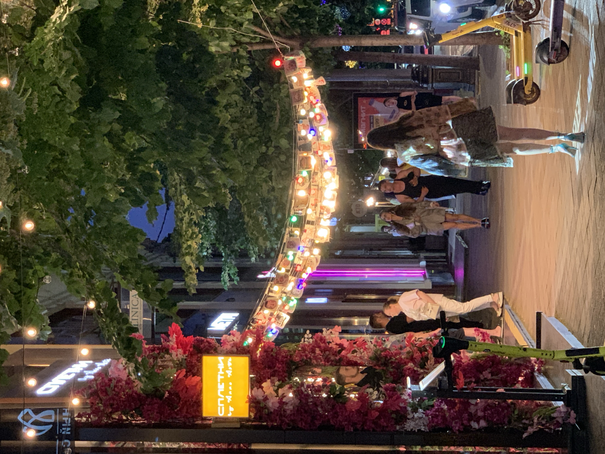 The image depicts an outdoor scene at night, likely on a street or pedestrian walkway in a city. A string of decorative lights, adorned with what appear to be small photos or cards, is strung across the path, illuminated by numerous small bulbs. Lush green trees overhang the area, their leaves silhouetted against the dusky sky.
On the left side of the scene, a vibrant display of pink and red flowers cascades down a structure. A yellow sign with white lettering is prominently displayed, reading "СПАЕТНИ by Anna Km". Beneath this sign, a man in a black shirt is partially visible.
Several people are walking along the paved walkway. In the foreground, a woman in a patterned jacket and a patterned skirt with light blue heels walks away from the camera. To her right, a yellow electric scooter is parked, with another scooter in front of it. Further down the path, other individuals are seen walking and standing, some interacting with each other. One woman in a light-colored outfit stands near a building entrance. A man in a black tank top and dark shorts is seen walking towards the viewer.
The environment appears to be a commercial area, with buildings and signage visible in the background. The overall atmosphere suggests a lively evening setting, possibly a restaurant or entertainment district.