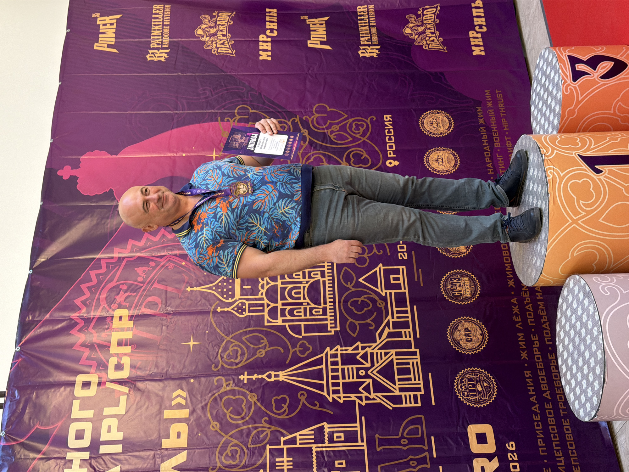 The image shows a man standing on a podium, holding a diploma and a medal. He is wearing a colorful, short-sleeved shirt and jeans. Behind him is a large purple banner with text and images in gold and pink. The banner features Cyrillic text, images of churches, and logos such as "POWER," "PAINKILLER HARDCORE DIVISION," and "DESPERADO." The podium has a metallic surface and is decorated with an orange and white pattern. Numbers "1" and "3" are visible on other podiums in the foreground. The man appears to be at an awards ceremony or competition. Based on the banner text, which includes "РОССИЯ" (Russia) and hints of historical imagery, the event is likely in Russia. Given the architectural style depicted on the banner, reminiscent of traditional Russian churches, and the mention of "SUZDAL" in other contexts, it is plausible that this event is taking place in the city of Suzdal, Russia, known for its historical churches and kremlin.