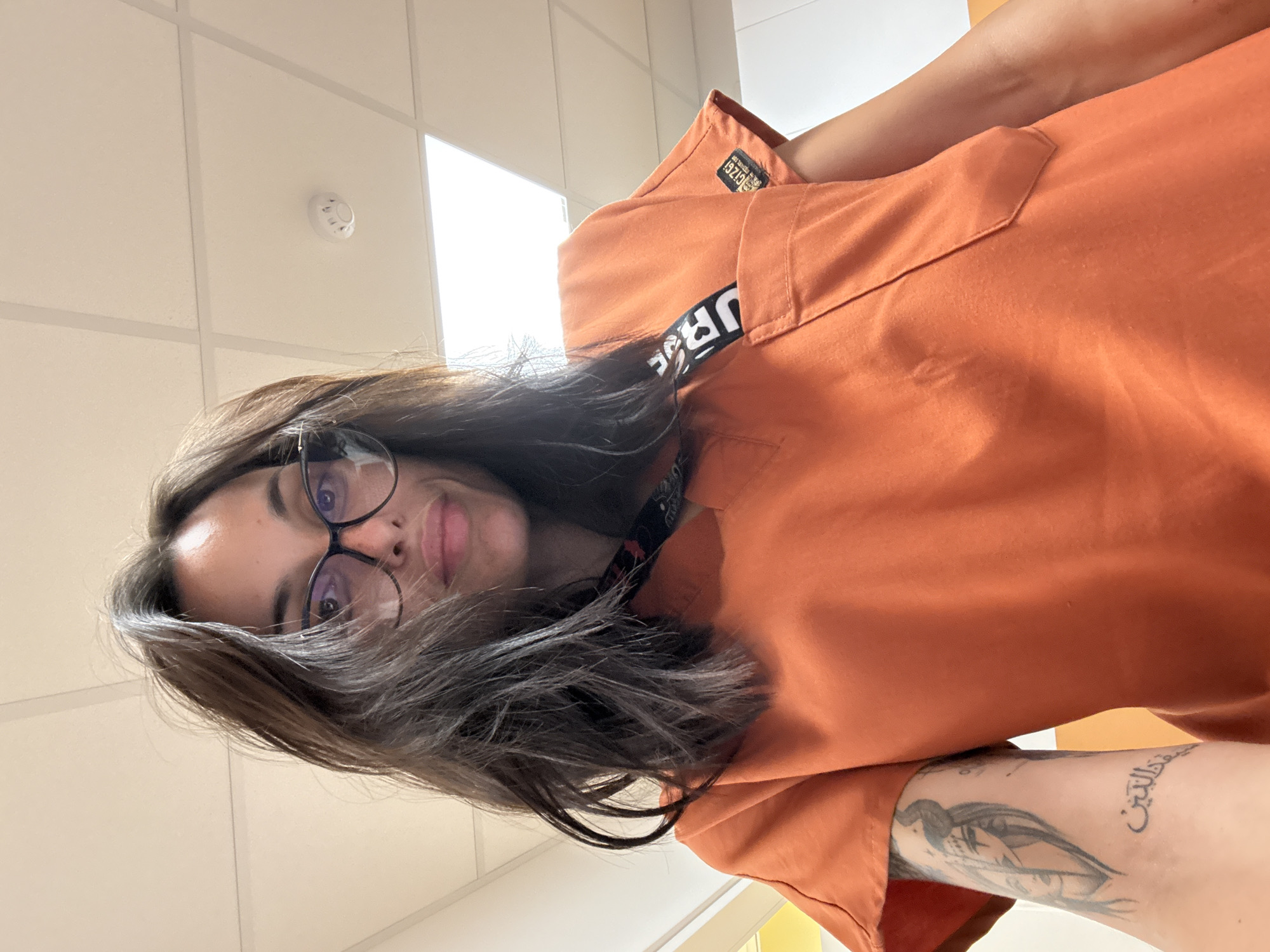 A woman is depicted in an upper-body portrait, captured from a low angle within an indoor commercial or institutional setting in Pleven, Bulgaria. She has dark, medium-length hair and wears black-framed eyeglasses. Her expression is neutral with a slight smile, and she looks directly at the camera. She is wearing an orange V-neck scrub-style top, with a black and white lanyard, partially displaying "URG," around her neck. A black patch with white text, including "GIZG1" and "urgnt.com," is visible on her right sleeve. Her left forearm features two tattoos: one depicting a stylized female face with a headdress or horns, and another of Arabic script below it. The background reveals a light-colored drop ceiling with rectangular acoustic tiles and overhead fluorescent panel lighting. A white circular smoke detector is affixed to the ceiling in the upper right. Light-colored walls are visible, with a faint yellow/orange element in the far left background.
