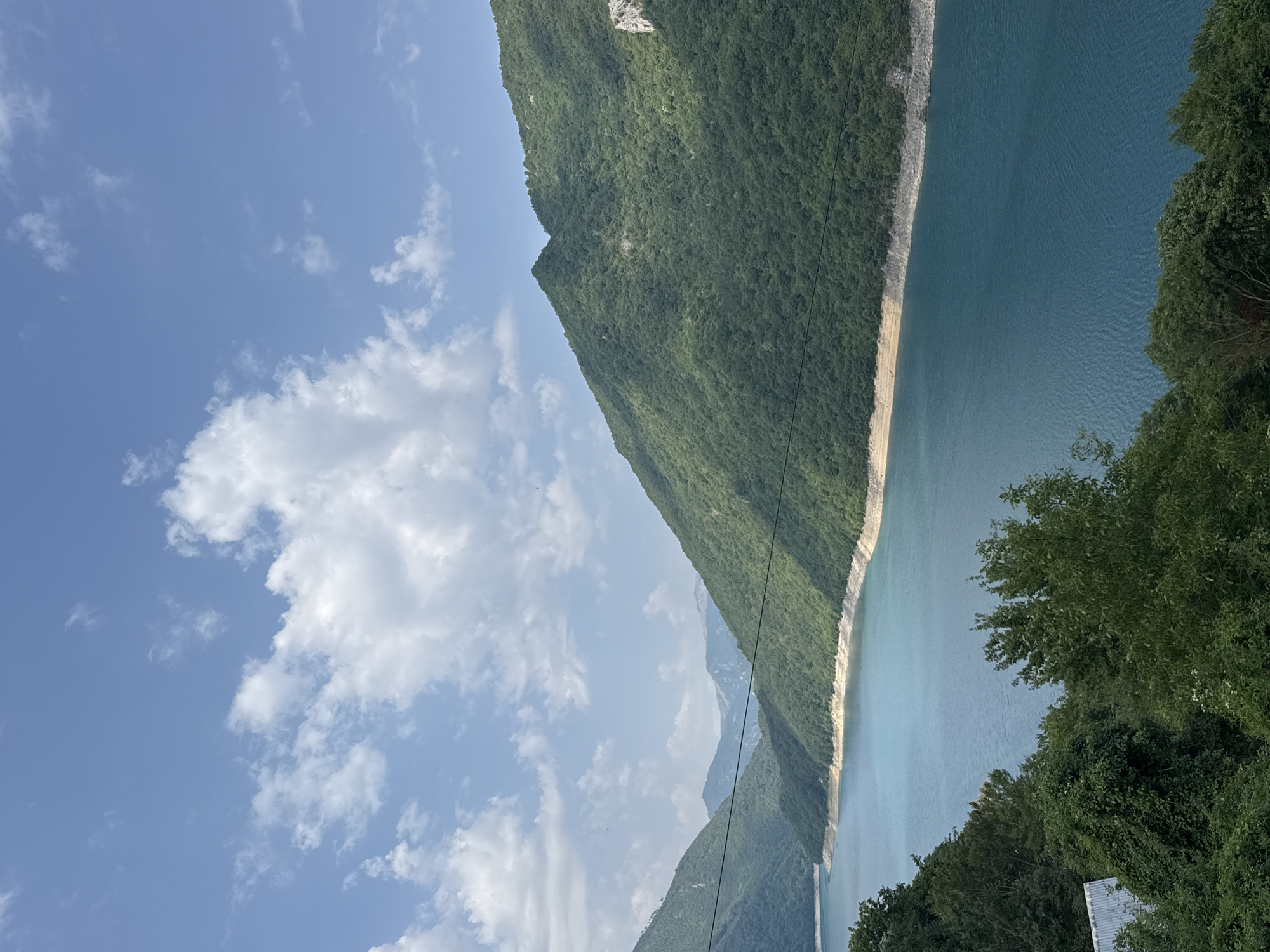The image depicts a wide shot of the Piva Lake in Plužine, Montenegro, under a partly cloudy blue sky. Lush green, tree-covered mountains flank the vibrant turquoise water, with a sandy shoreline visible along the base of the mountains. The foreground is filled with dense green foliage, obscuring a small portion of a white structure with a corrugated roof on the left. Two thin black lines, possibly wires, stretch horizontally across the frame. The lighting suggests it is daytime, with shadows cast on the mountains, indicating the sun is positioned to the left of the frame.