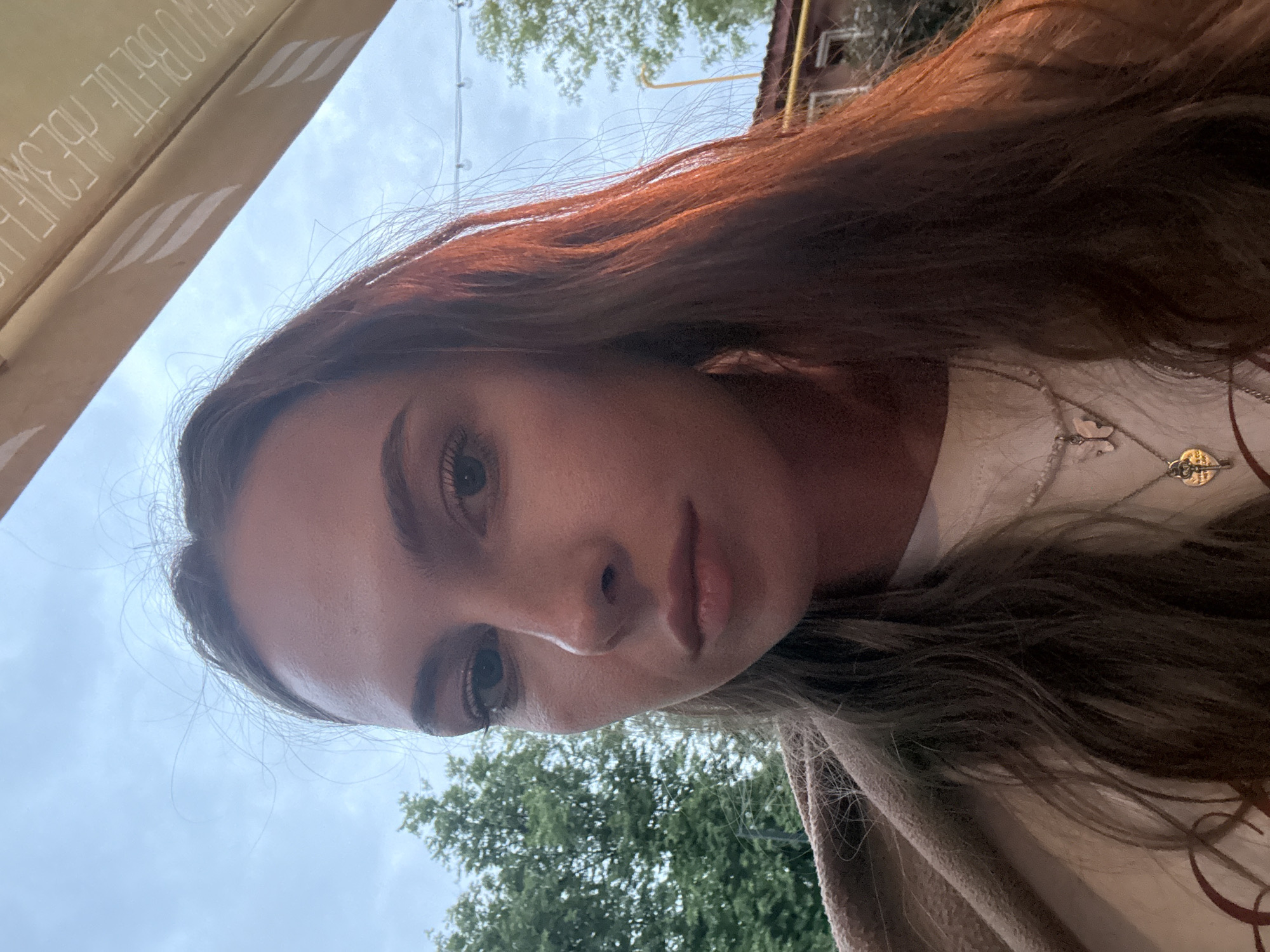 A young woman is depicted in a close-up, upward-angled outdoor portrait in Perm, Russia. Her face occupies the central and upper-left portion of the frame, with her gaze directed forward, meeting the camera. She has light skin, prominent eyelashes, and full lips. Her long, dark brown hair flows down her shoulders, displaying reddish highlights on the right side due to lighting. She wears a light-colored top and two layered necklaces: one featuring a silver-toned butterfly pendant and another with a heart-shaped key pendant. A light brown, textured fabric is draped over her left shoulder. The background features a partially overcast sky with green tree foliage visible on the left and upper-left. On the upper-right, an angled, light-colored awning or canopy is visible, bearing inverted text and a striped pattern. In the lower-right background, part of a building with a reddish roof, yellow piping, and string lights can be discerned. The scene is illuminated by natural outdoor light.