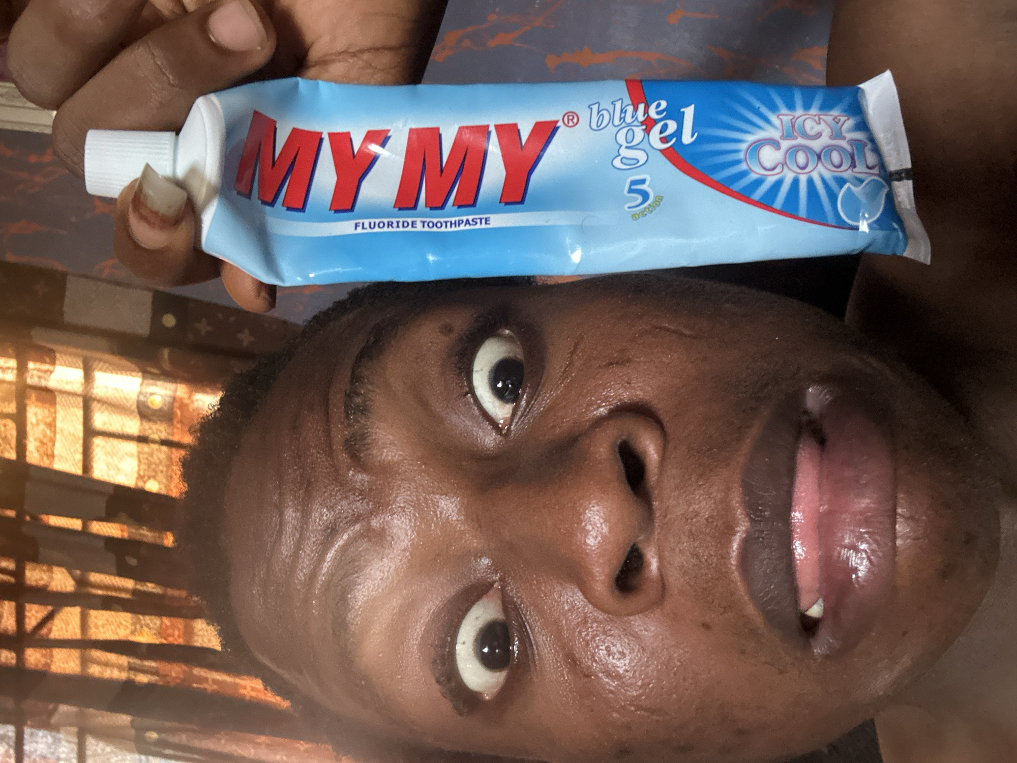 A person is holding a tube of "MY MY Blue Gel Icy Cool" fluoride toothpaste. The person is facing the camera with a slightly open mouth and wide eyes. The background appears to be a room with a window. The location is Unknown, Unknown.