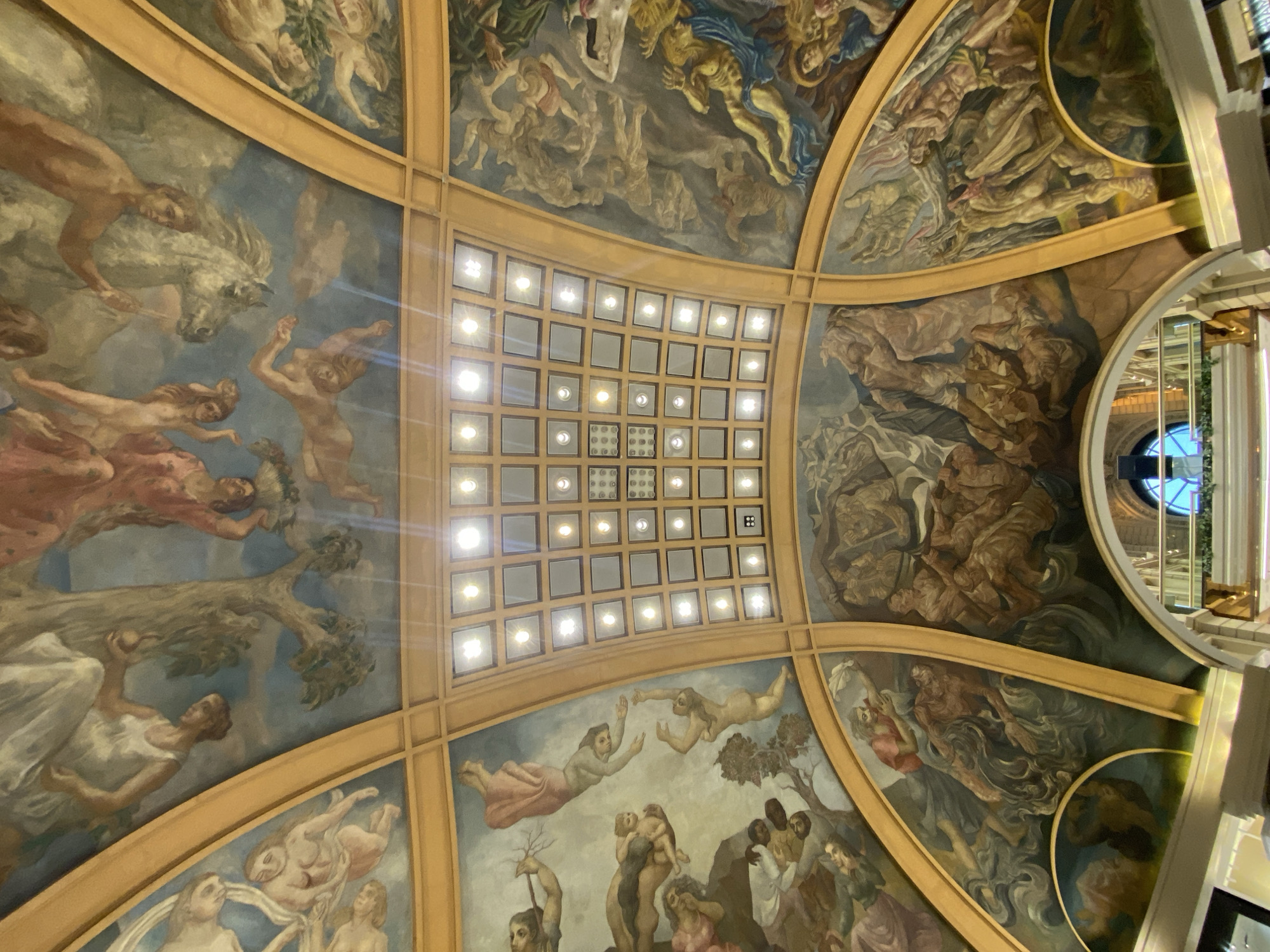 The image shows a view of a painted ceiling with a grid of square lights in the center. The paintings depict various figures and scenes in classical style, with a focus on mythological or allegorical subjects. The figures appear to be predominantly nude or draped in flowing garments, and include humans, horses, and possibly other animals. The paintings are separated by a gold-colored frame. The overall palette consists of muted earth tones, blues, and whites. In the upper right corner, a round window and architectural details are visible. This appears to be a section of the dome of Galerías Pacífico, a shopping mall in Buenos Aires, Argentina, known for its ornate architecture and ceiling murals.