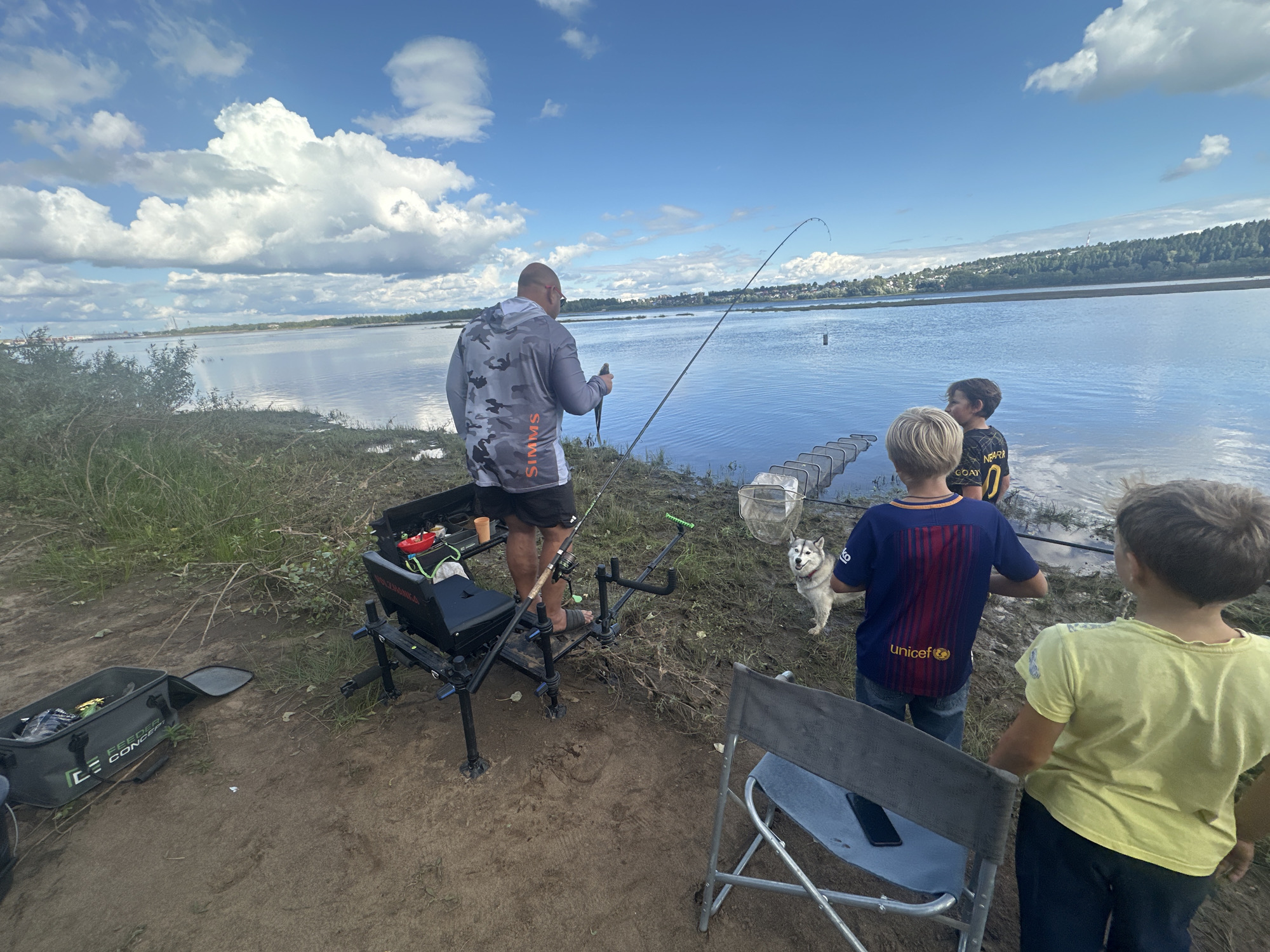 The image captures a group of people fishing by a large body of water, likely a river or lake, on a cloudy day. In the foreground, a man in a camouflage hoodie holds a fish and a fishing rod. To his right, two young boys stand near a husky dog. One boy wears a blue soccer jersey, and the other a yellow t-shirt. Several fishing rods, nets, and equipment are visible around them. The background shows a distant treeline and buildings, suggesting a semi-urban or suburban environment. The ground is muddy and covered with grass. The location is identified as Noviy, Russia, based on the image's metadata.