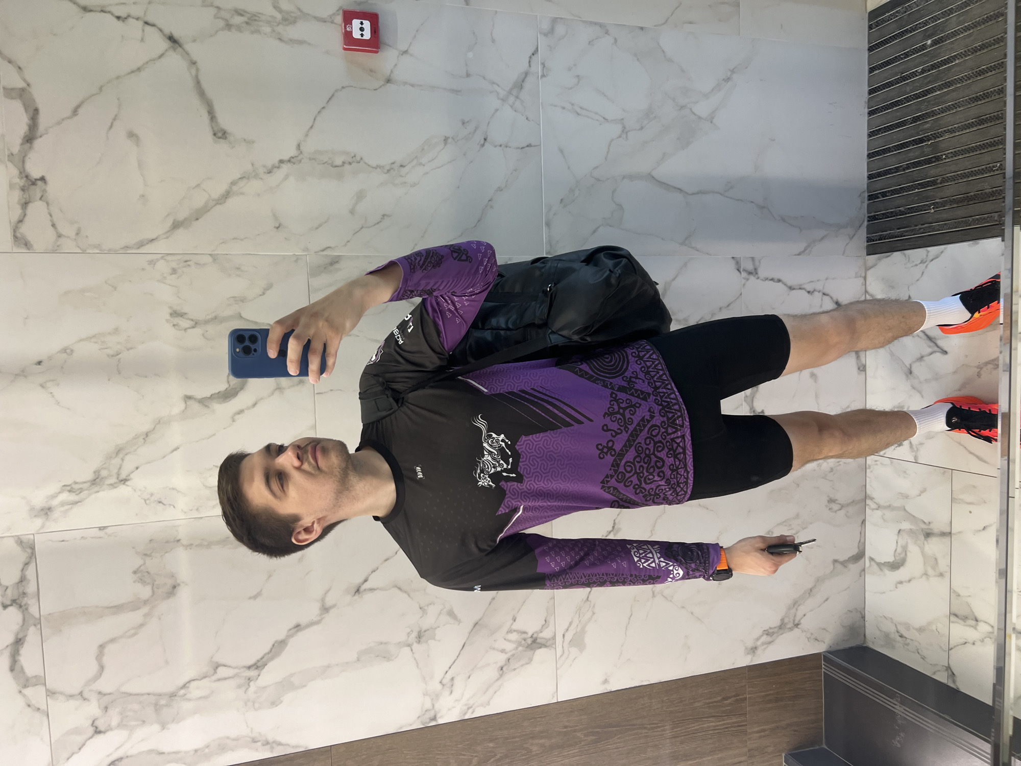 A person is standing in what appears to be a bathroom or restroom, indicated by the marble tile walls and the partially visible mirror on the right side. The person is holding a blue smartphone in their left hand, seemingly taking a selfie. They are wearing a black and purple long-sleeved shirt with patterns, black shorts, white socks, and orange and black running shoes. A black duffel bag is slung over their shoulder. They are also holding a key in their right hand. The wall has a red emergency button. The location is suspected to be in Kazan, Russia.