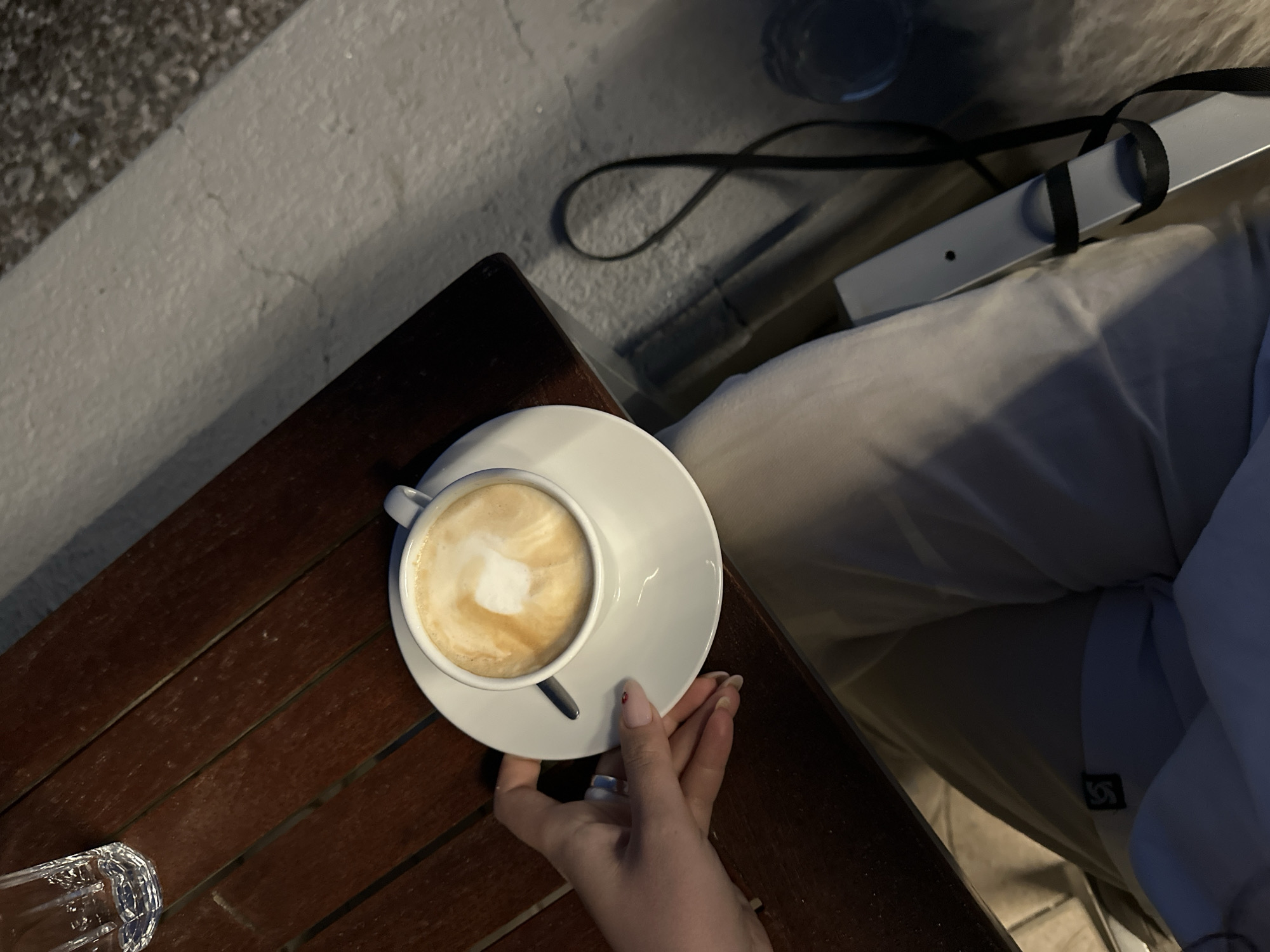 The image shows a top-down view of a table with a cup of cappuccino on a saucer. A hand is holding the saucer. A glass of water is also on the table. Part of a person wearing light-colored pants is visible in the background, along with what appears to be a black strap. The location is implied to be Šušanj, Montenegro, but there are no visual indicators in the image to confirm this.