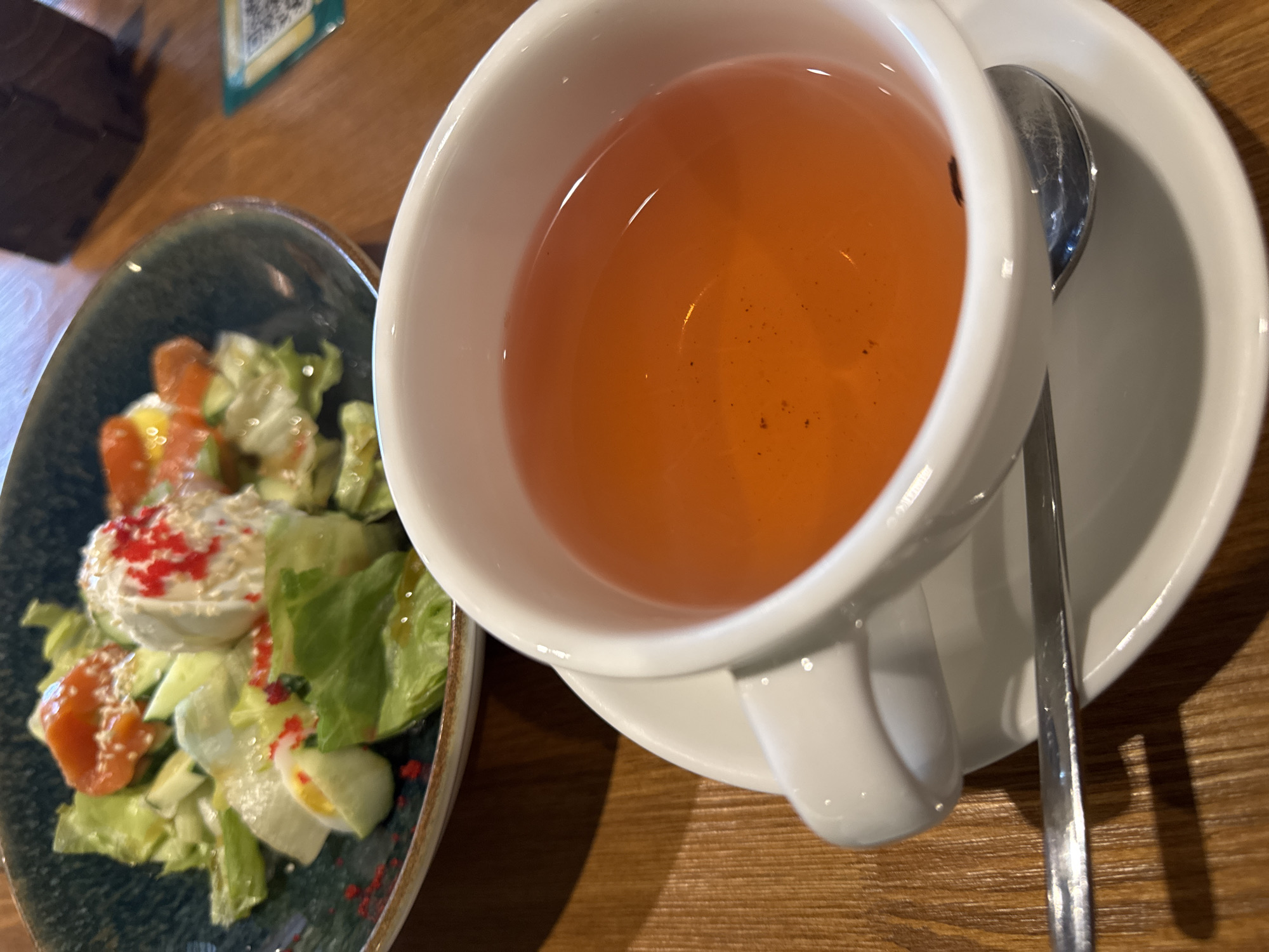 A close-up shot shows a white cup of tea and a salad on a wooden table. The tea is a warm, amber color, with a few dark specks visible at the bottom of the cup. The cup sits on a matching white saucer, and a silver spoon rests on the saucer. In the background, a salad is served in a dark blue bowl. The salad appears to be made of lettuce, sliced eggs, and pieces of salmon, garnished with red caviar and sesame seeds. The scene is lit by natural light, creating soft shadows on the wooden table. The image was taken in Kazan, Russia.